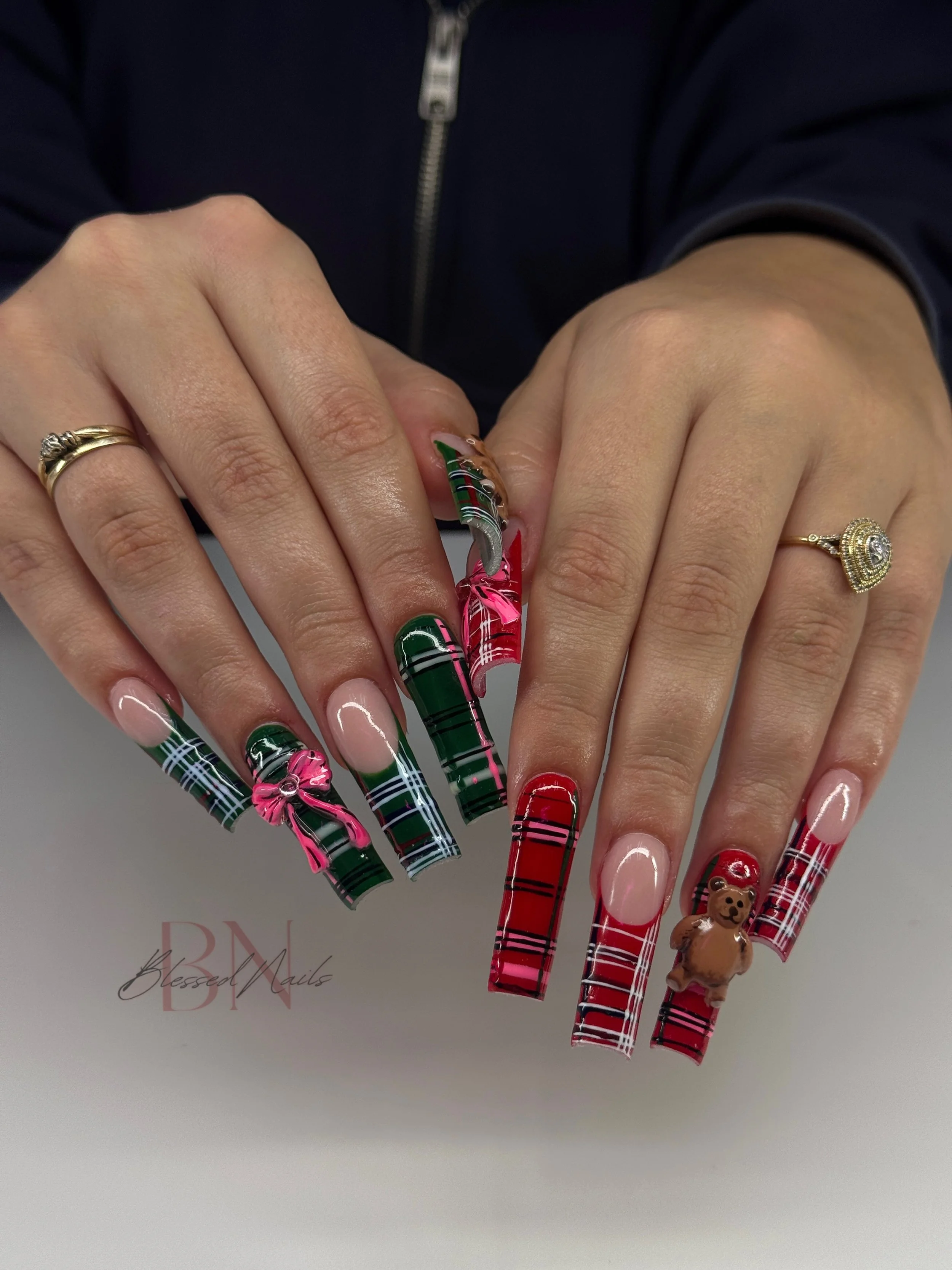 Close-up of hands with Christmas-themed nail art featuring red, green, and white plaid designs, pink bows, a teddy bear, and a holly leaf. Hands have rings and are positioned over a white surface.