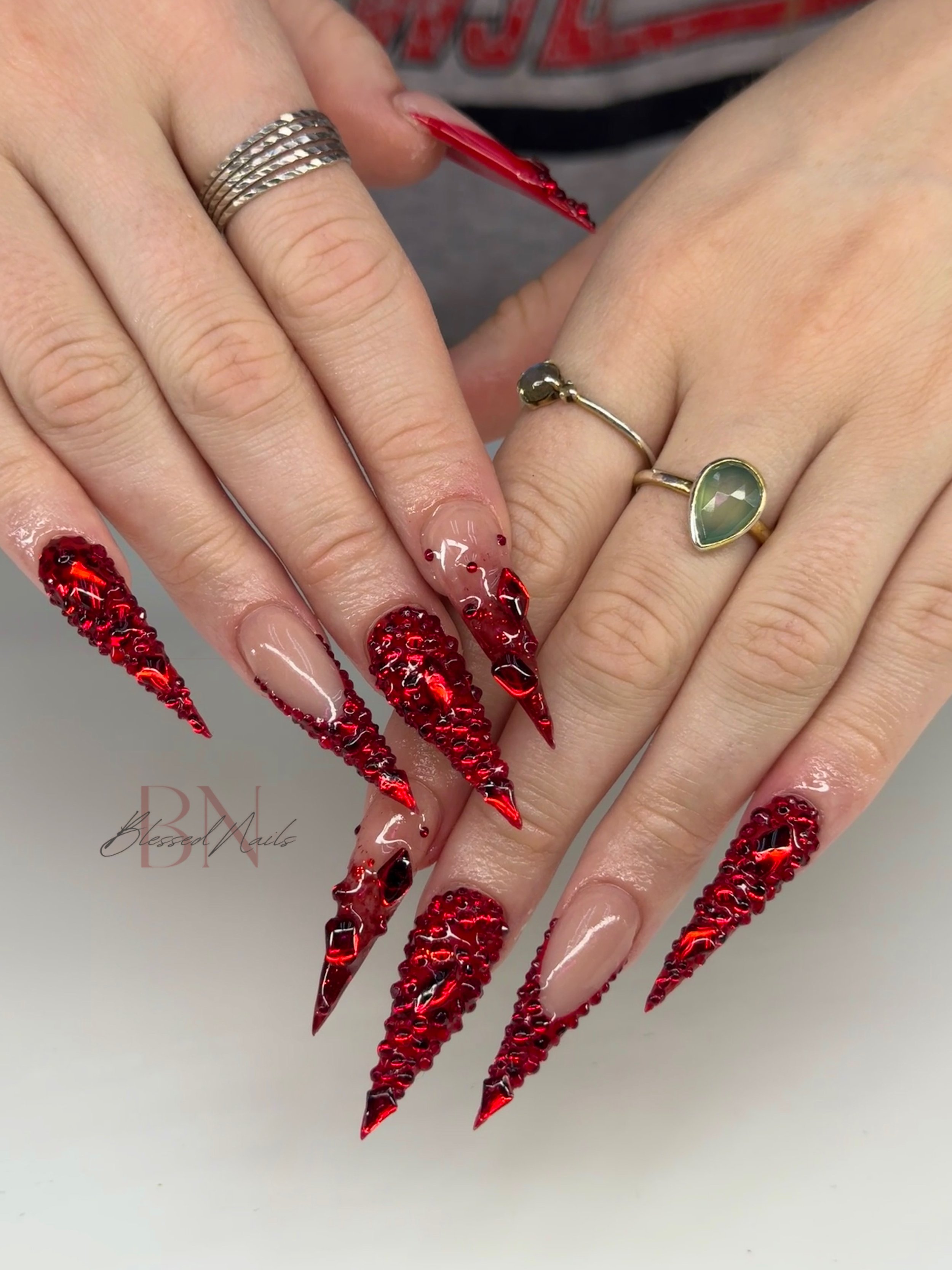 Close-up of hands with long, stiletto-shaped acrylic nails in a red, textured, 3D design with black accents, wearing multiple rings including a silver band and a green gemstone ring.