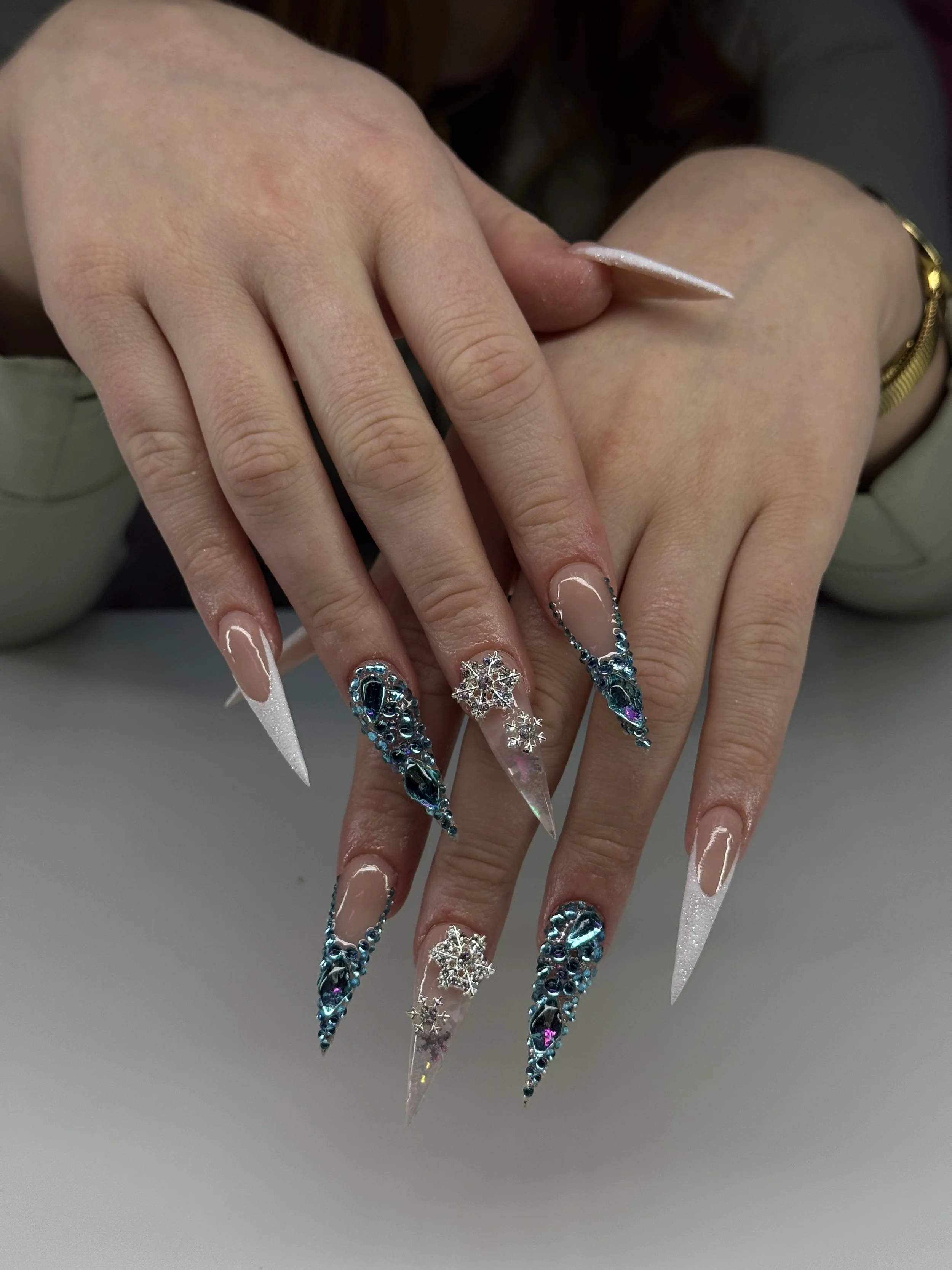 Close-up of a woman's hands with long, decorated acrylic nails and rings, resting on a white surface.