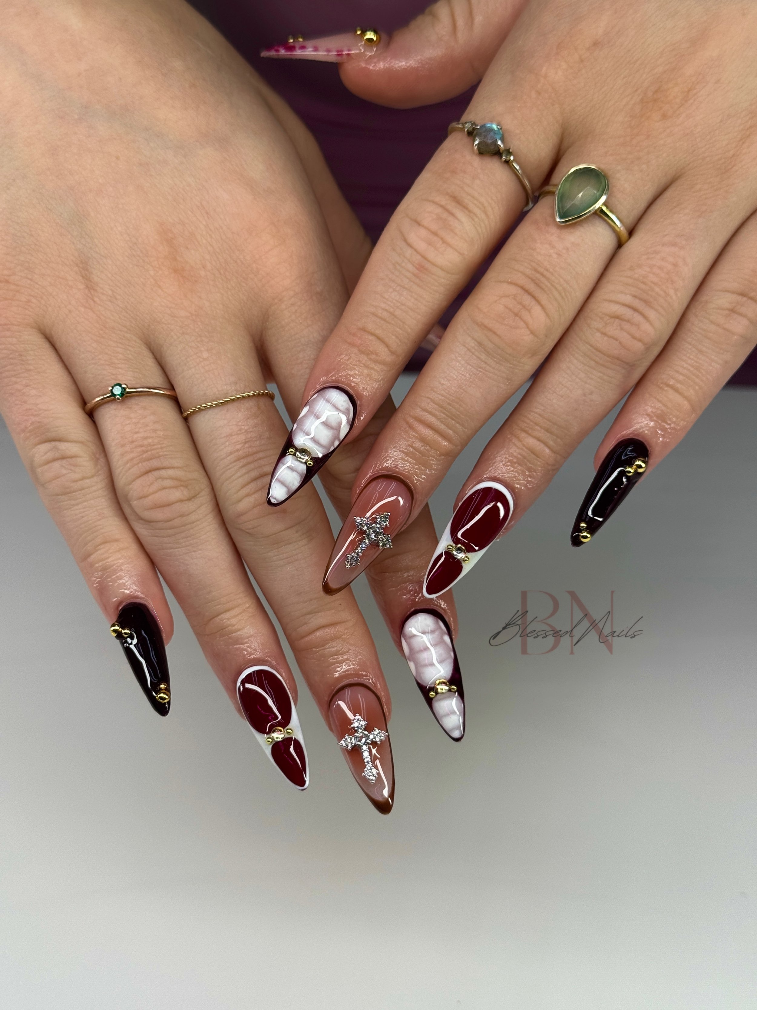 Close-up of a person's hands with decorated long stiletto nails and multiple rings, including a cross ring and various gemstone rings, against a light background.