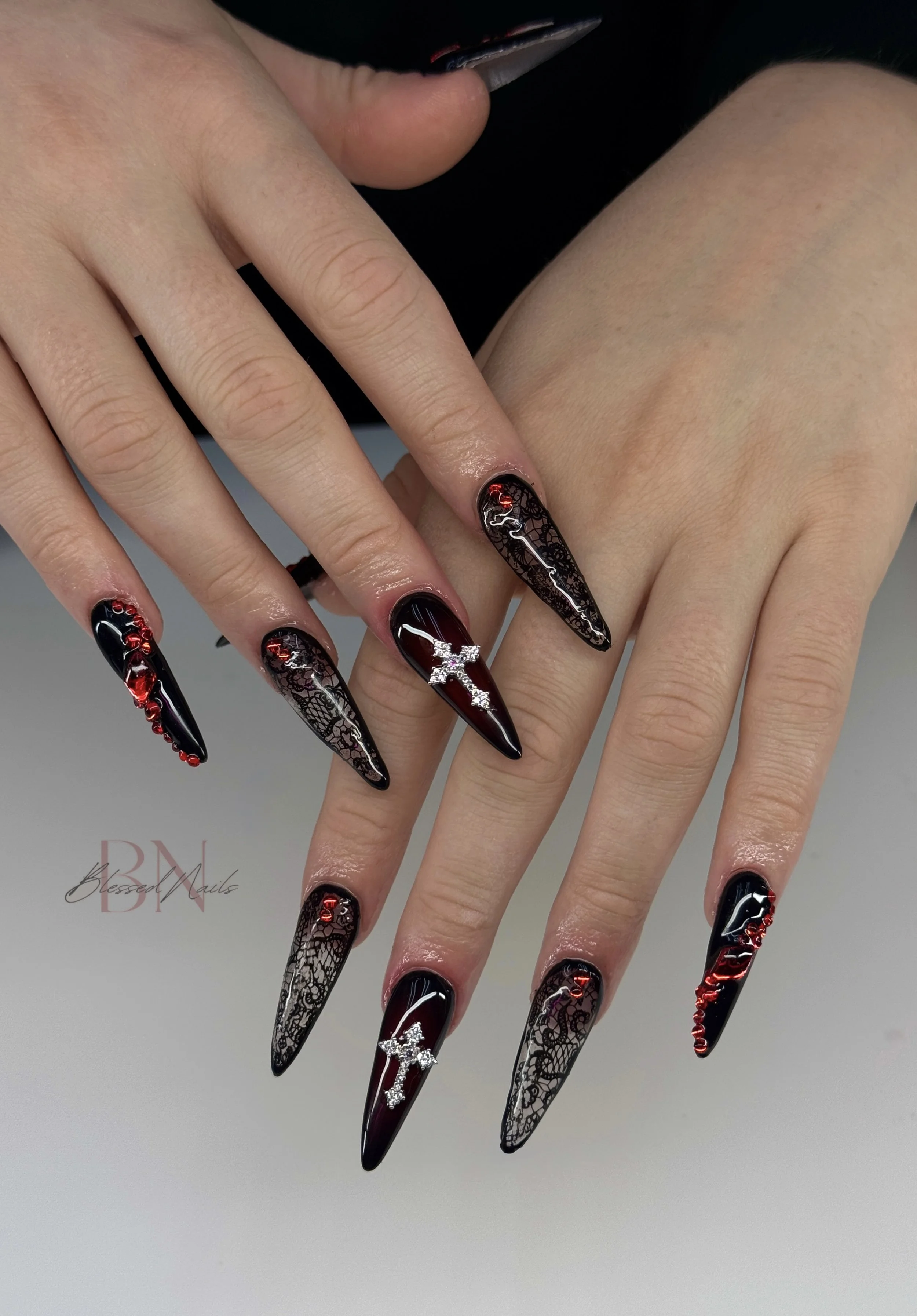 Manicured nails with black, red, and lace design, decorated with rhinestones and cross charms.