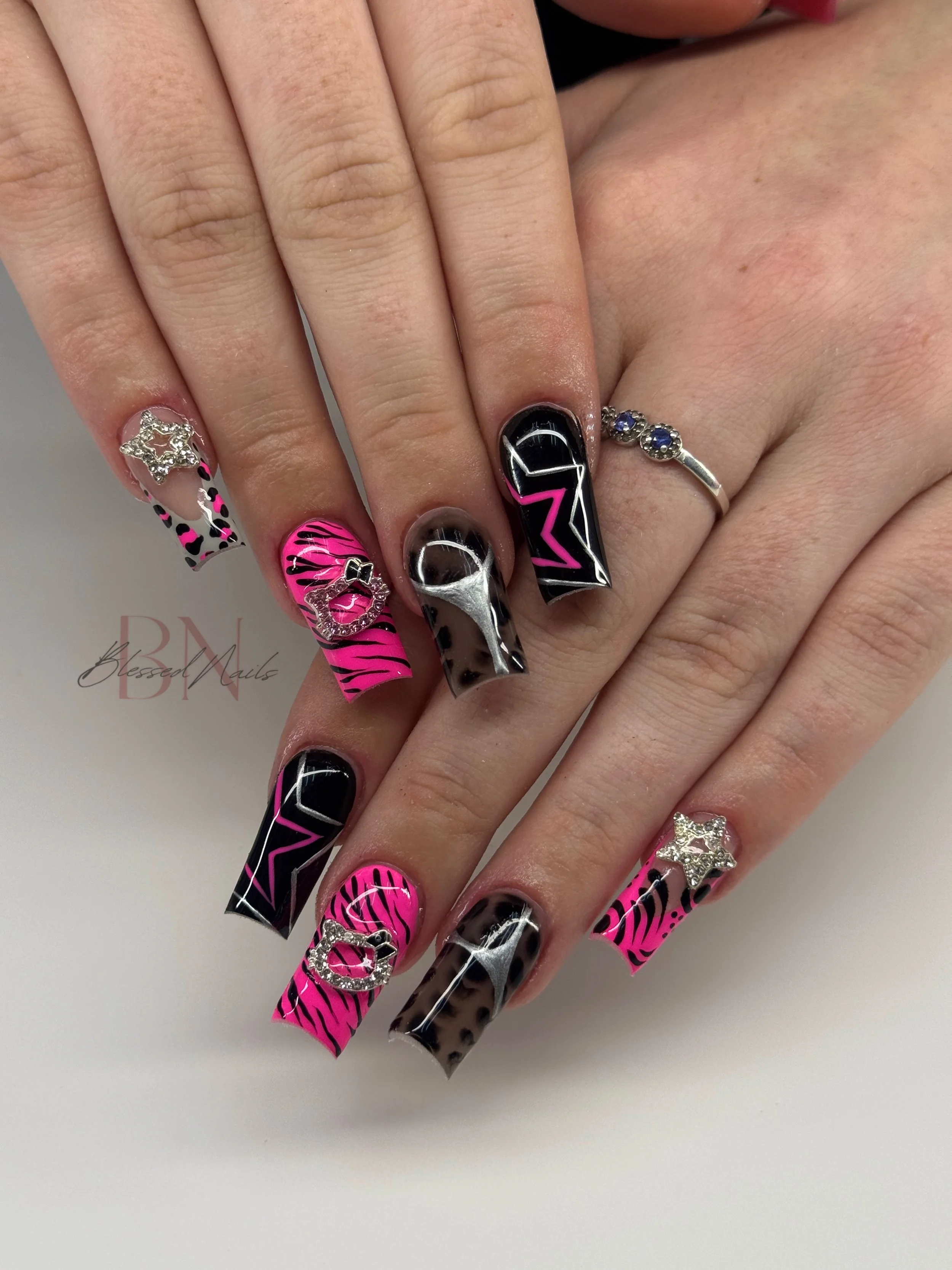 Close-up of hands with decorated black and pink animal print and geometric patterned nails, some nails have rhinestone bows or star charms, and one has a silver heart charm.