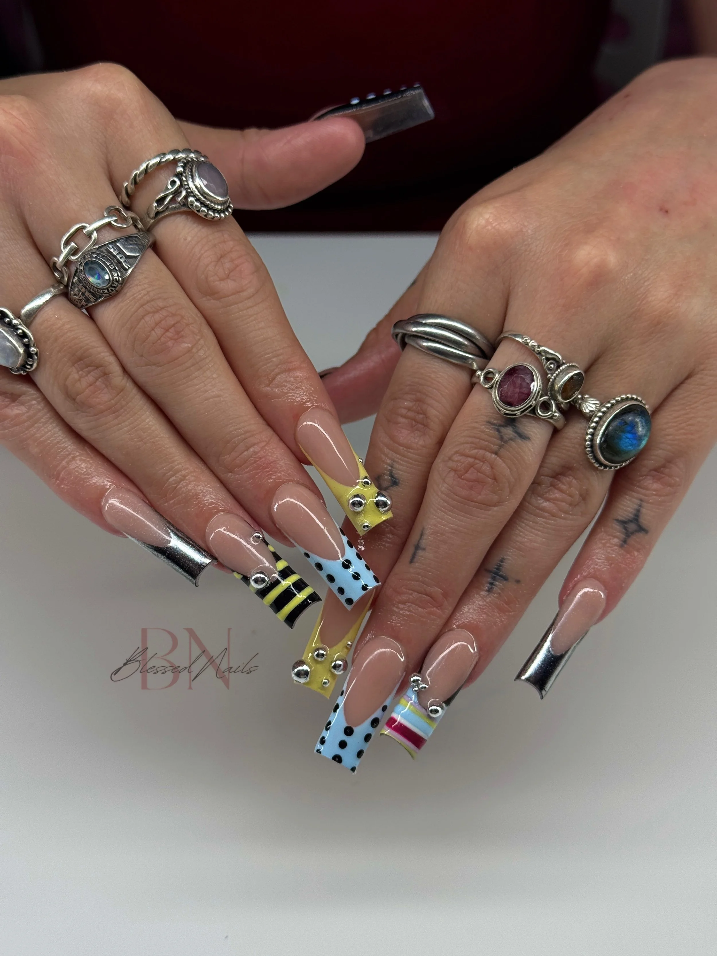 Close-up of a person's hands showing colorful, patterned long acrylic nails, adorned with silver rings and hand tattoos, against a white background.
