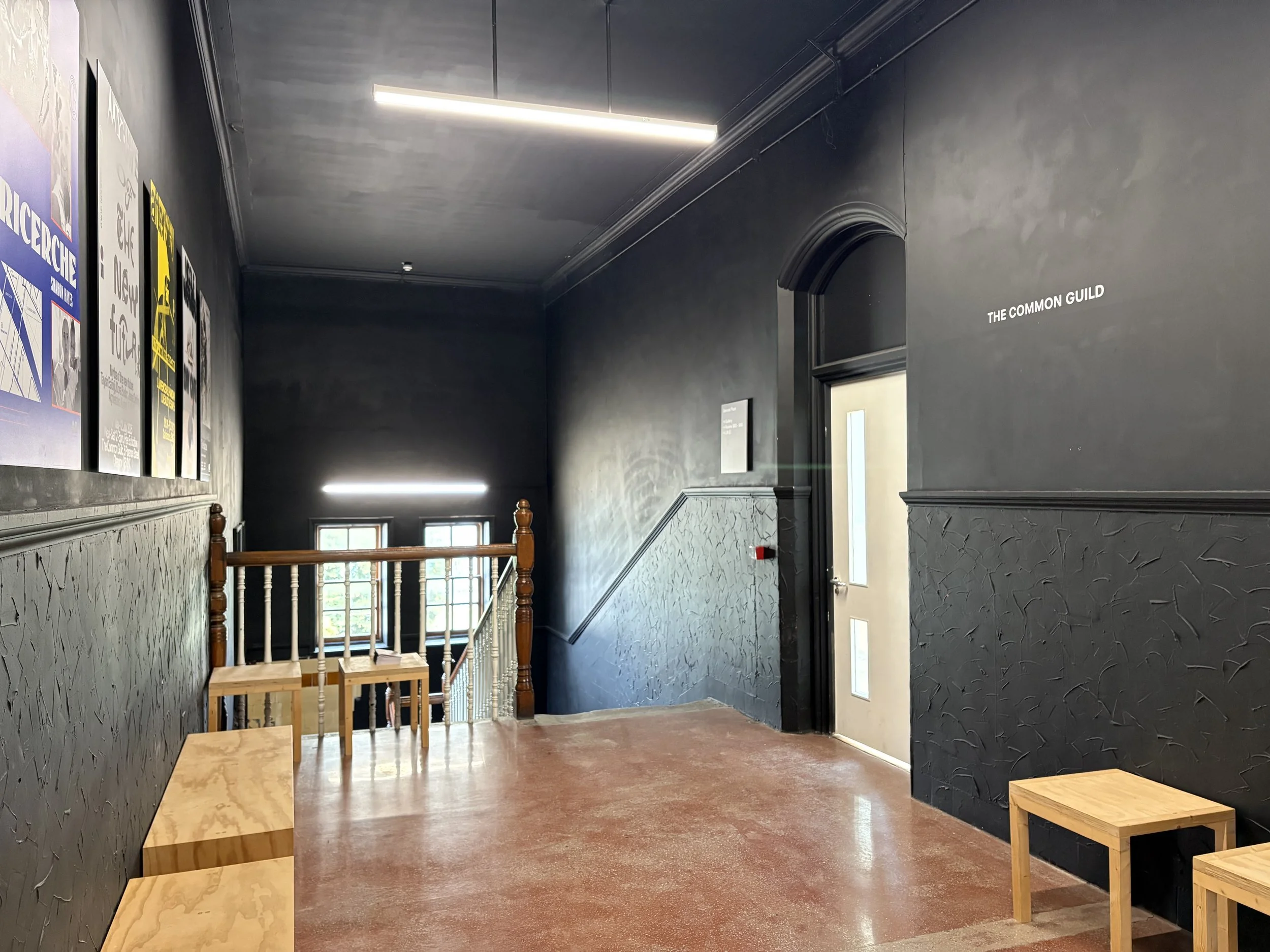 A landing painted in black with a row of low wooden benches on its left and a white door on its right. Five posters hang on the left-hand wall.