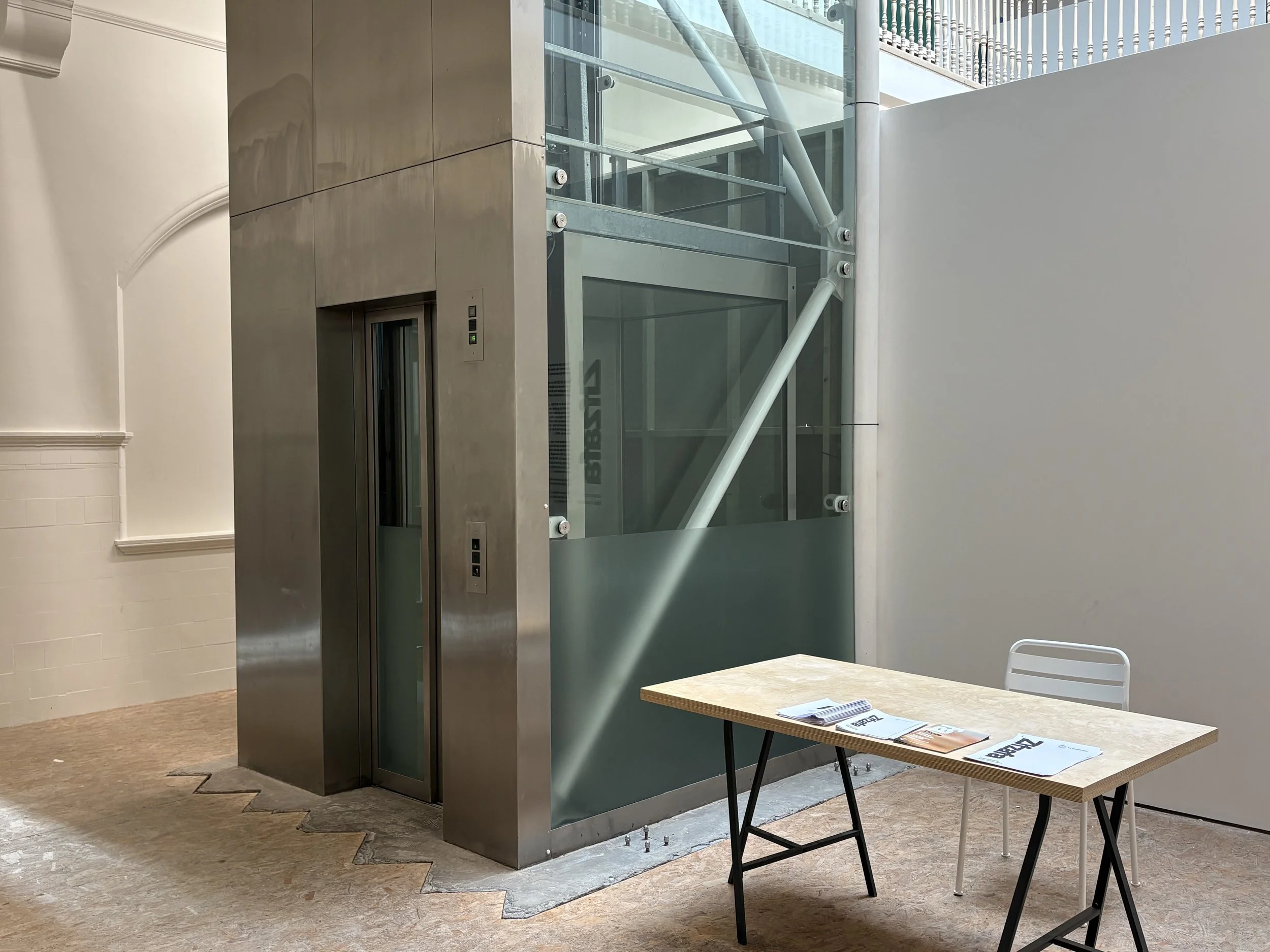 A steel and glass lift in the centre of the ground floor in an atrium. A table and seat for an invigilator stand to the right of the lift entrance.