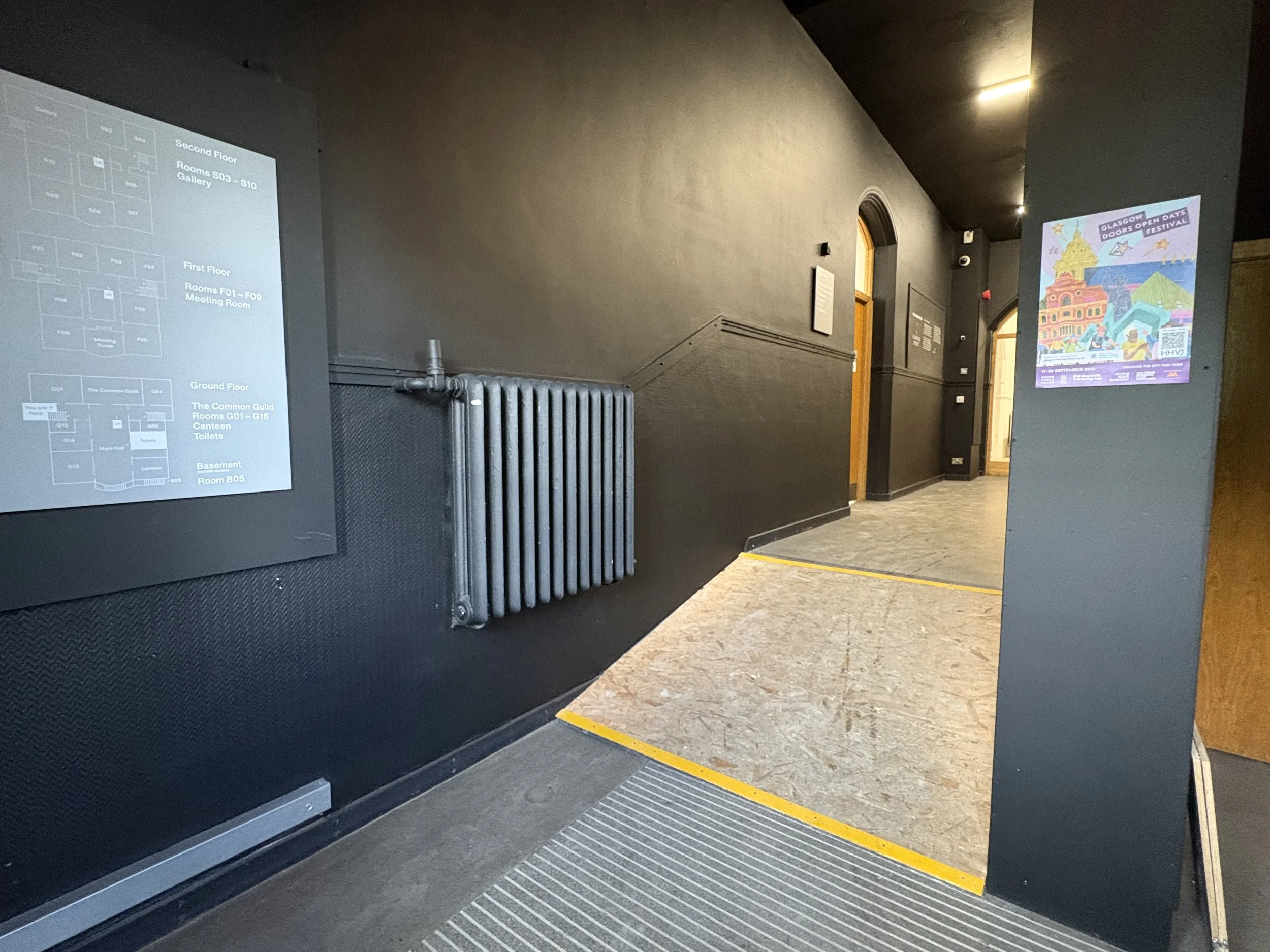 A corridor painted with walls painted in black leads to a wooden ramp which leads on to an atrium space. A plan of the building hangs on the left wall.