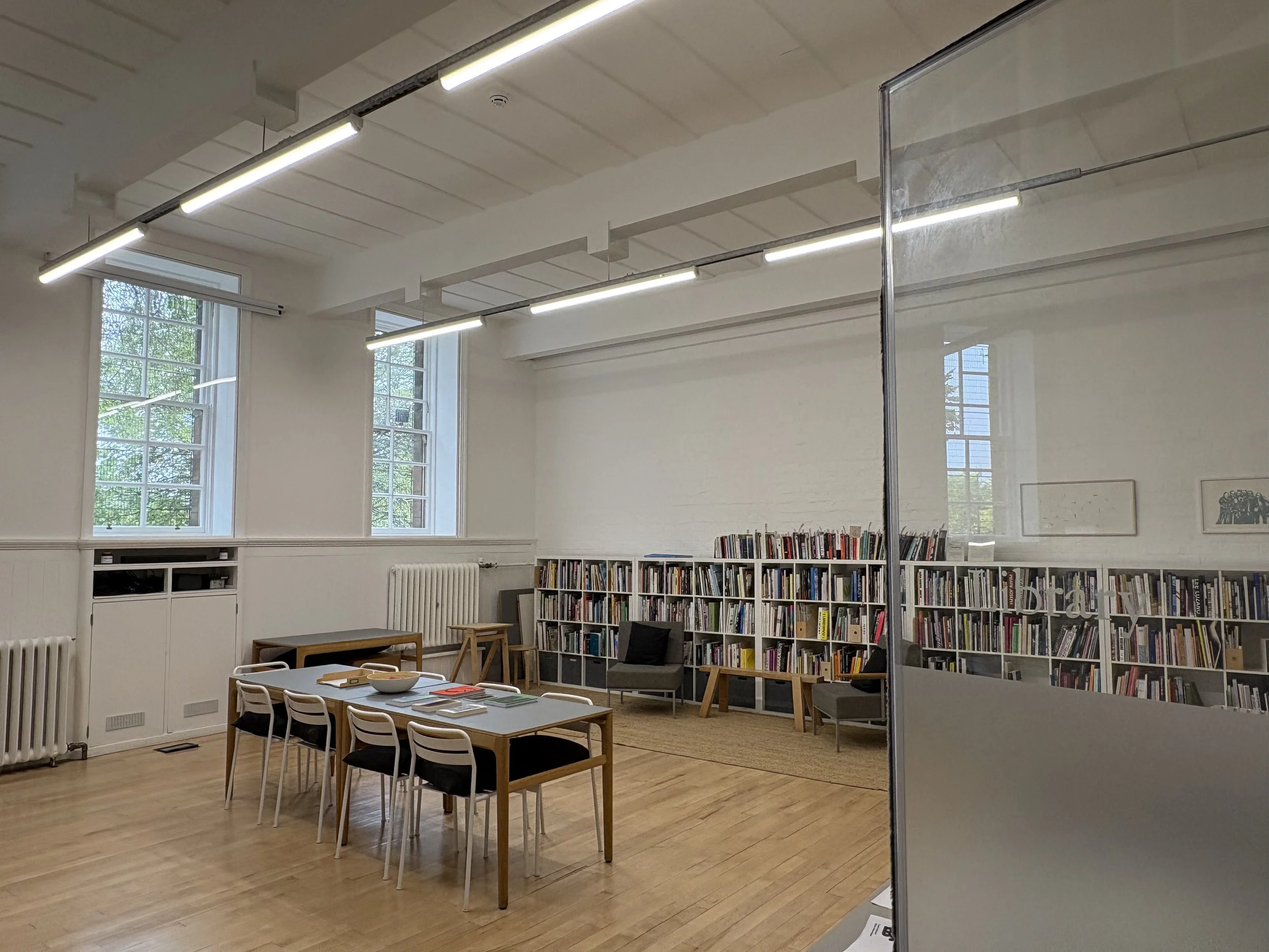 A large room painted in white with two large windows and wooden floors. Along the back of wall runs a long bookshelf and in the centre of the room is a table and chairs.