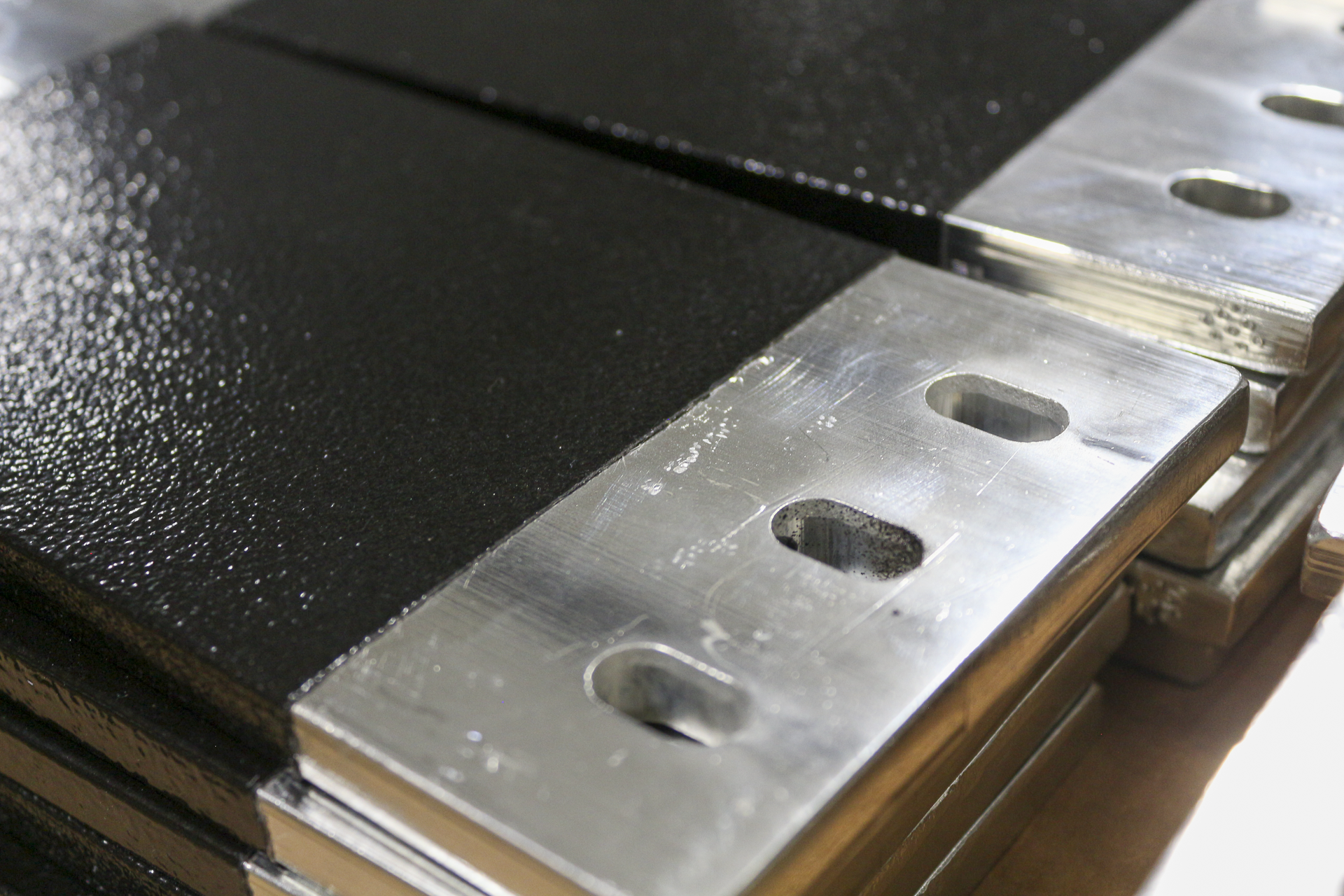 Close-up of a metal hinge attached to a black textured surface, with multiple elongated holes in the hinge.