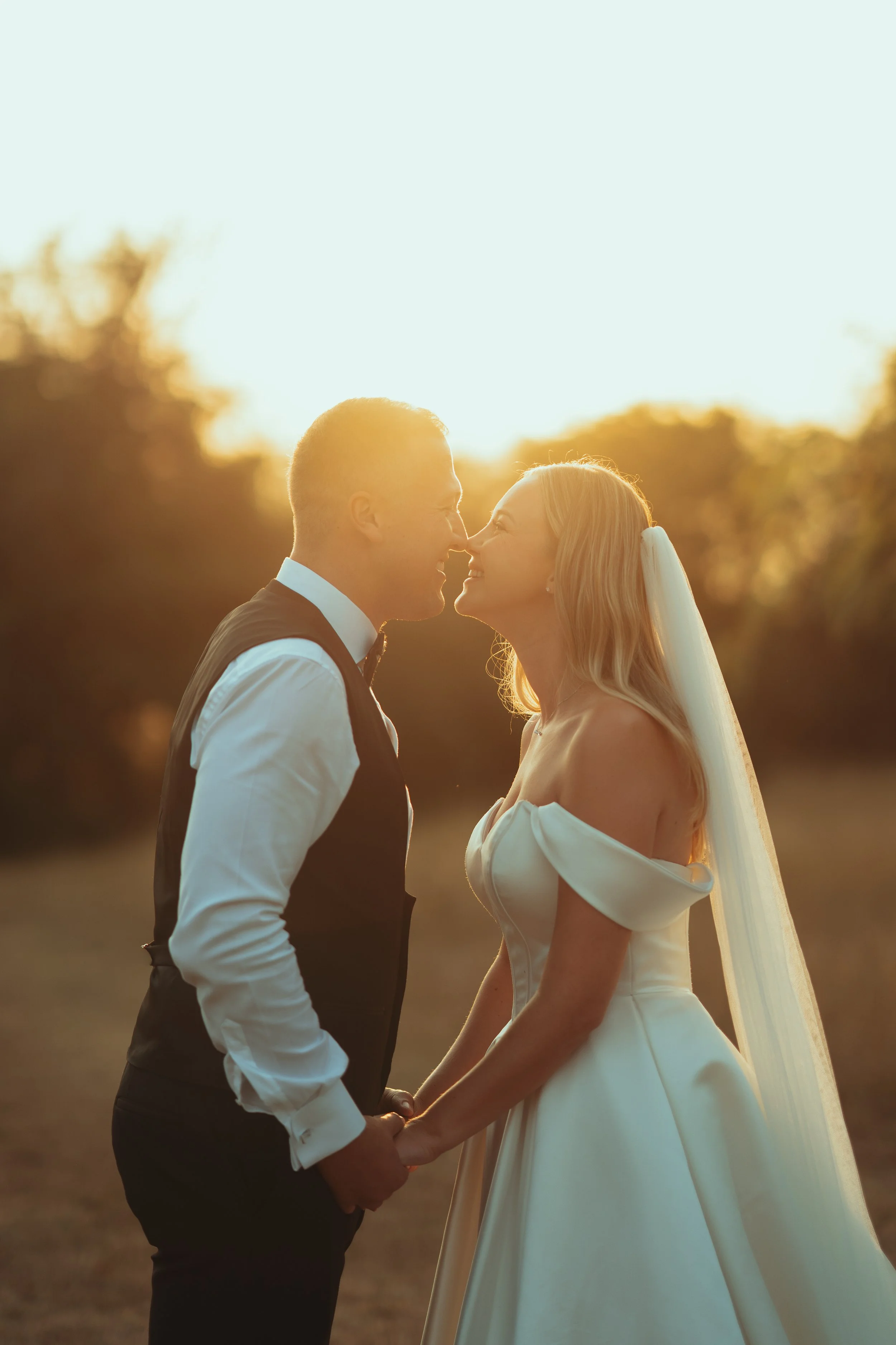 An engaged couple holding hands during their wedding at sunset, standing face-to-face closely, with a blurred natural background and the sun setting behind them.