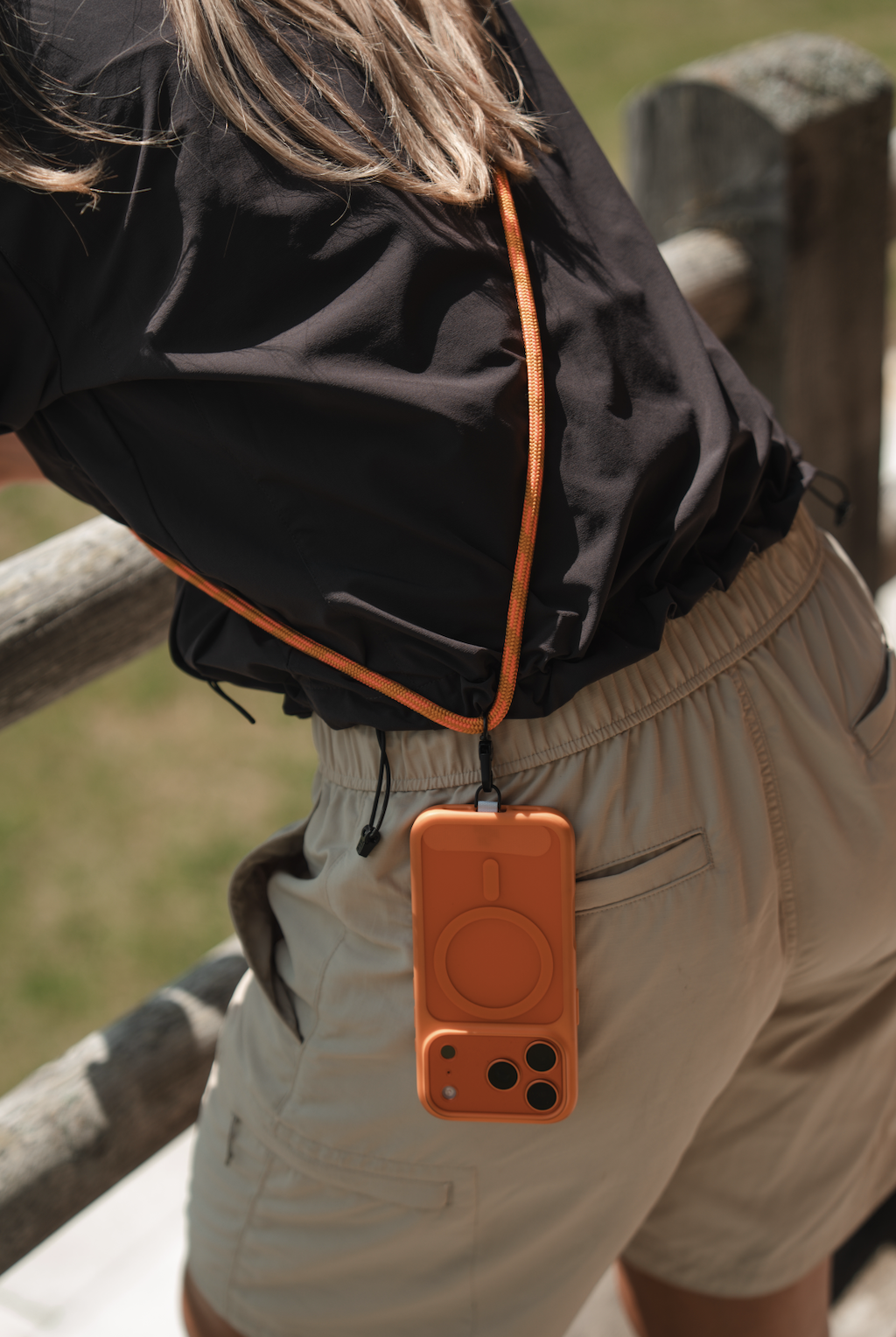 Person standing outdoors wearing beige shorts, black jacket, and an orange portable speaker hanging from a lanyard around their neck.
