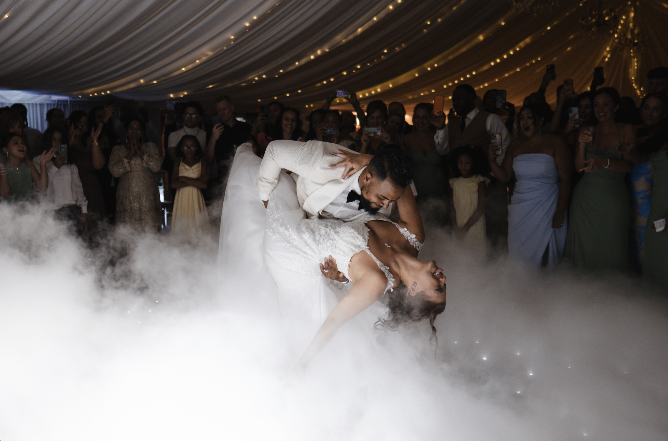 A bride and groom dancing at their wedding reception with guests watching and taking photos. The scene is illuminated with warm lighting and has a fog or smoke effect on the floor.
