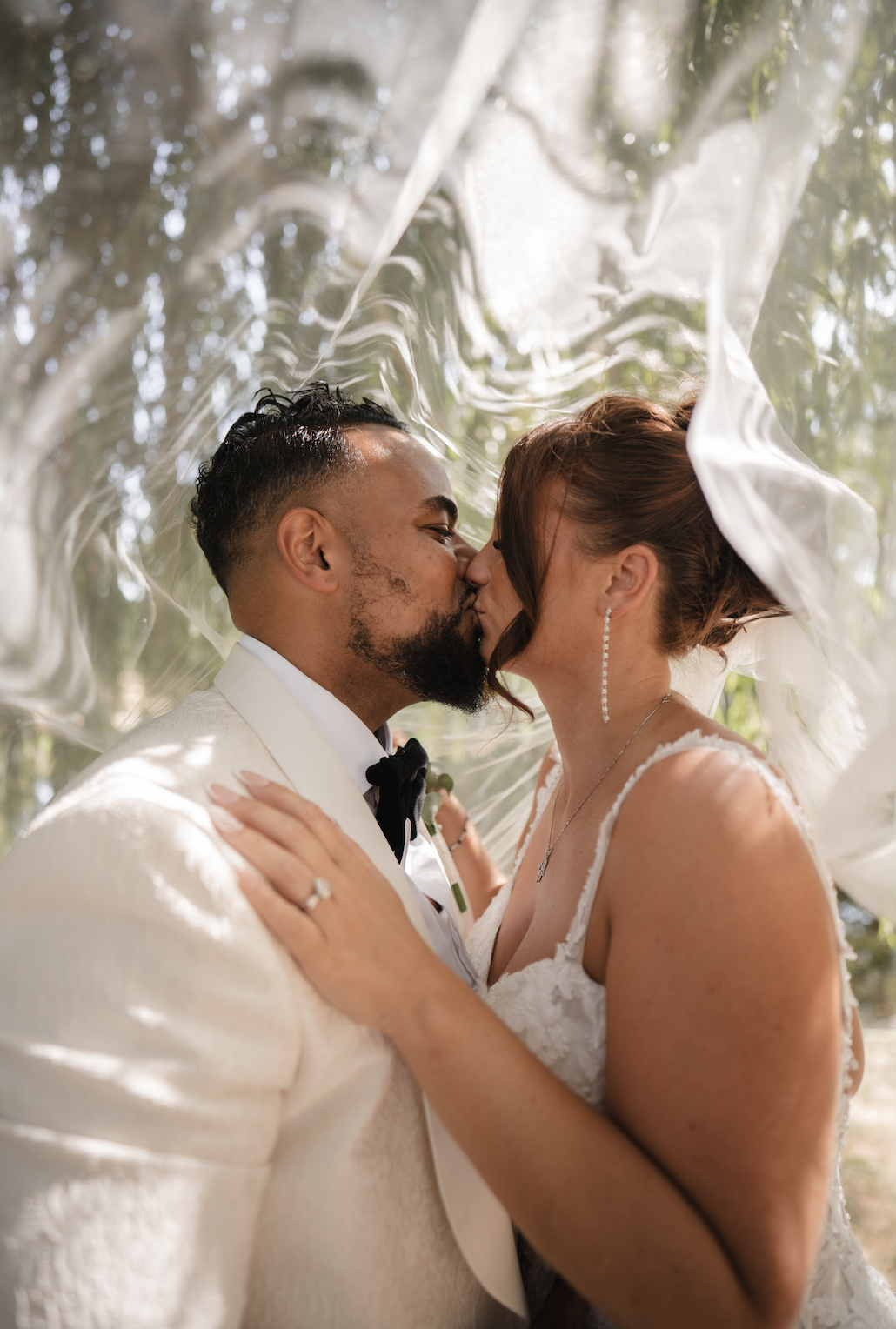 A newlywed couple sharing a kiss during their wedding ceremony, with sunlight filtering through trees in the background.