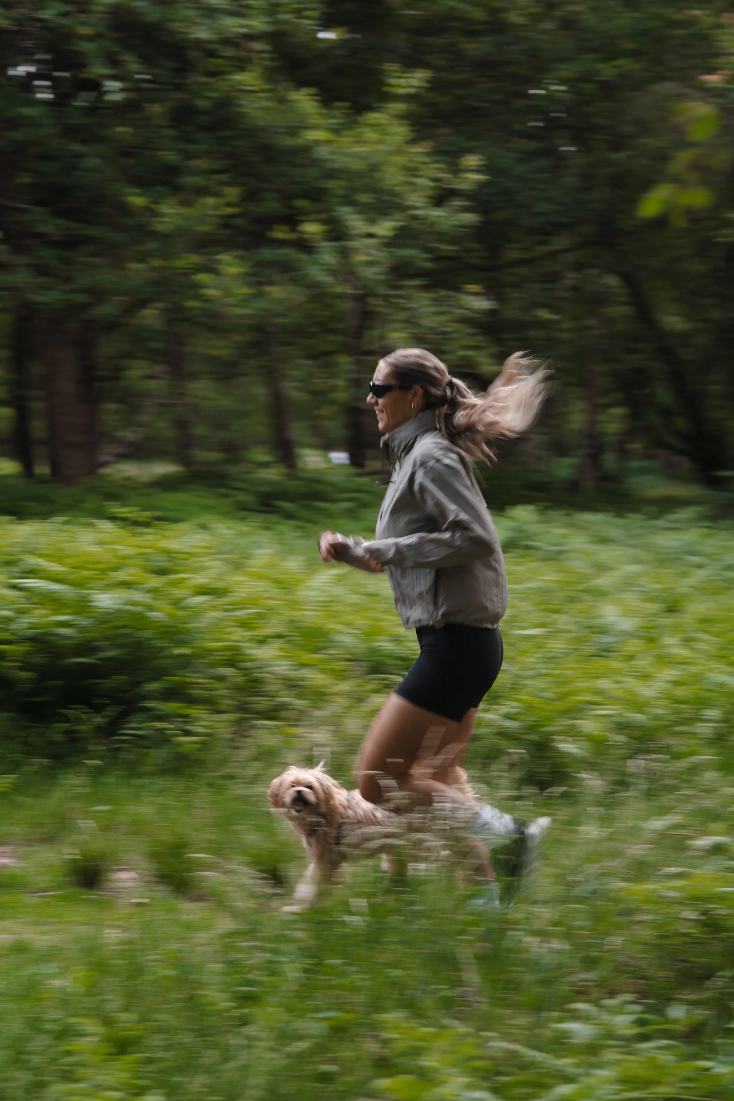 A woman wearing sunglasses, a gray jacket, and black shorts is jogging in a green forested area with a small brown and white dog running alongside her.