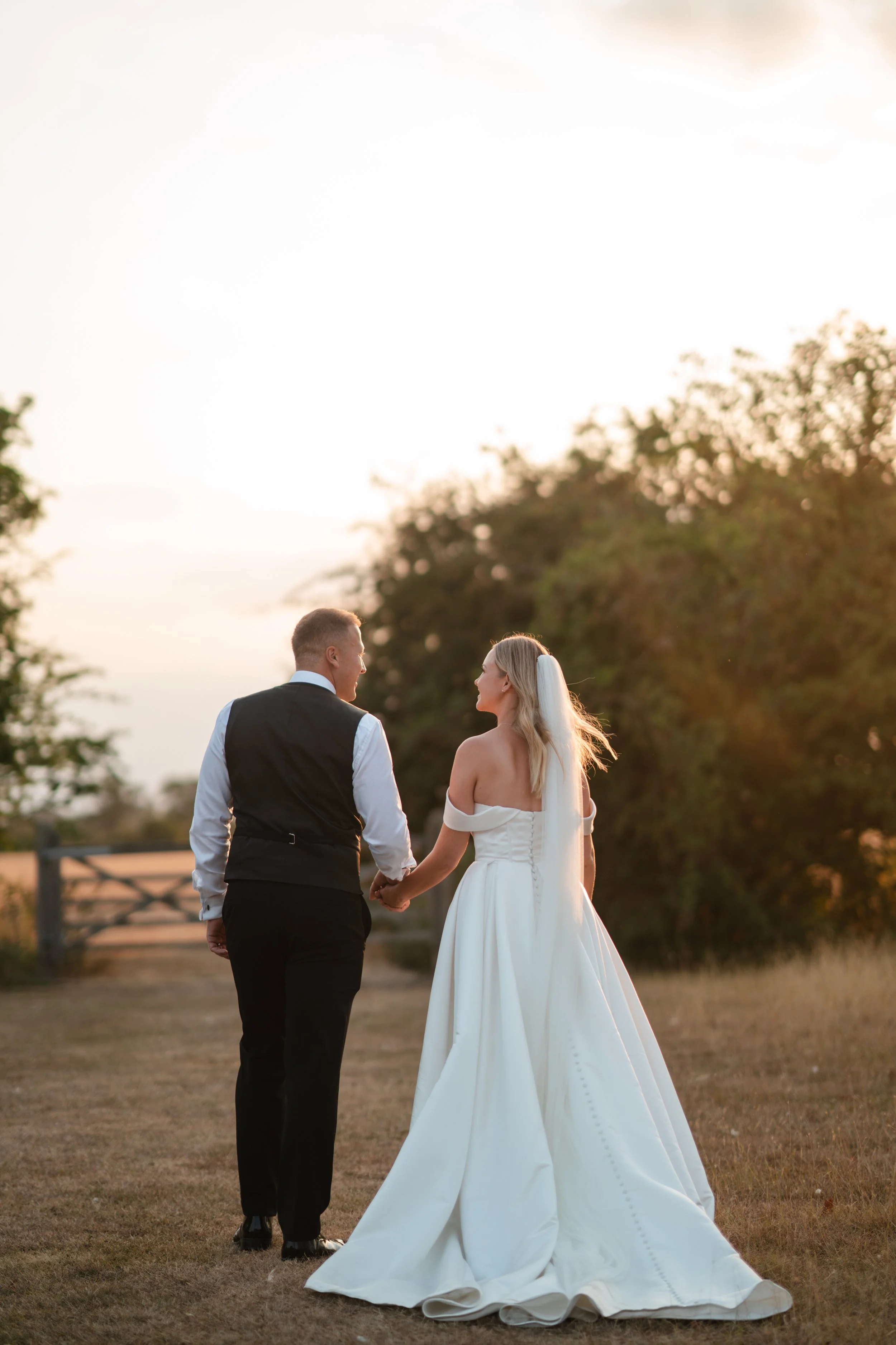 A bride and groom holding hands outdoors during sunset. The bride is wearing a white off-shoulder wedding gown with a long train, and the groom is dressed in a black vest, white shirt, and black pants.