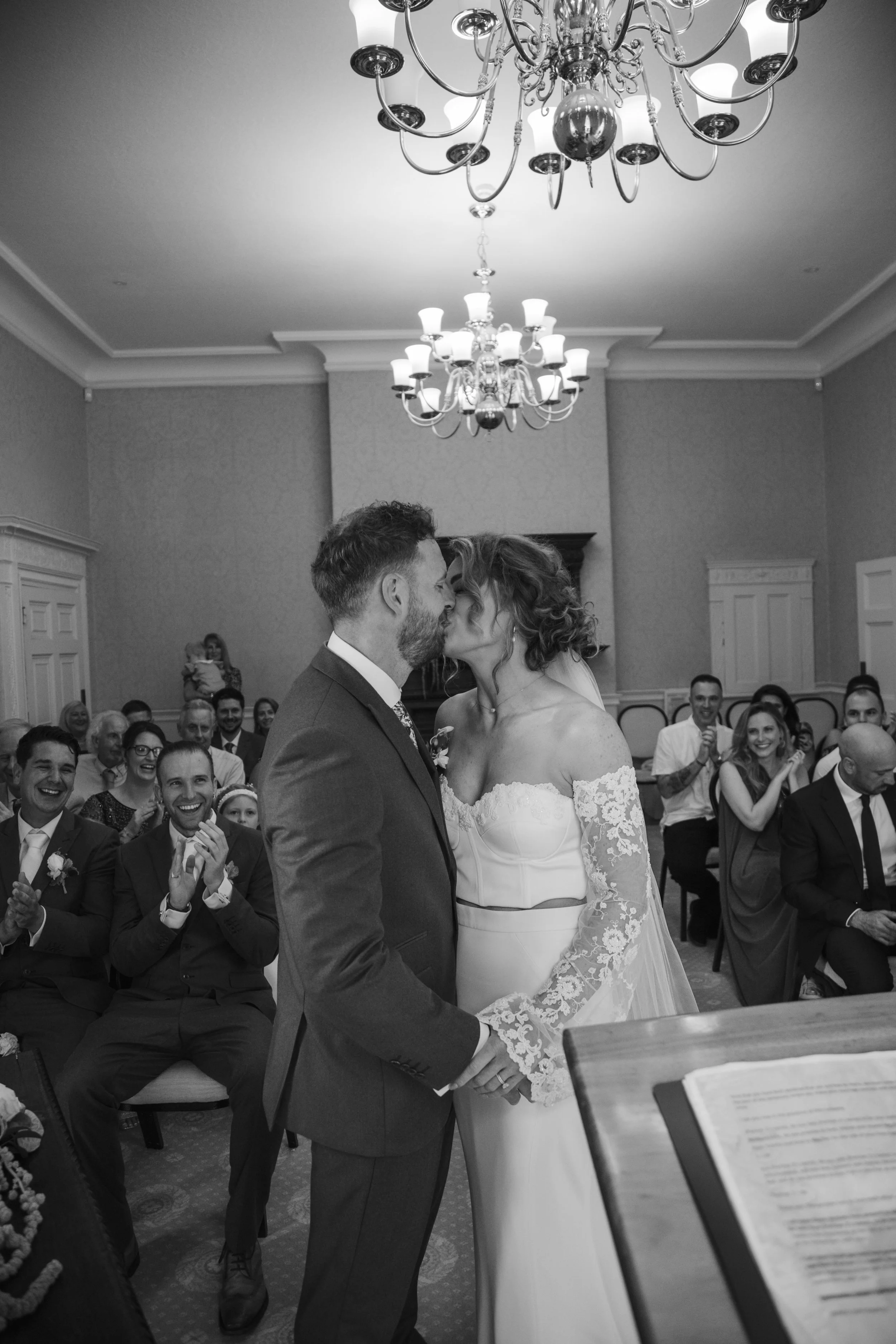 A black and white photo of a wedding ceremony with a bride and groom facing each other, surrounded by seated guests in a decorated room with chandeliers.