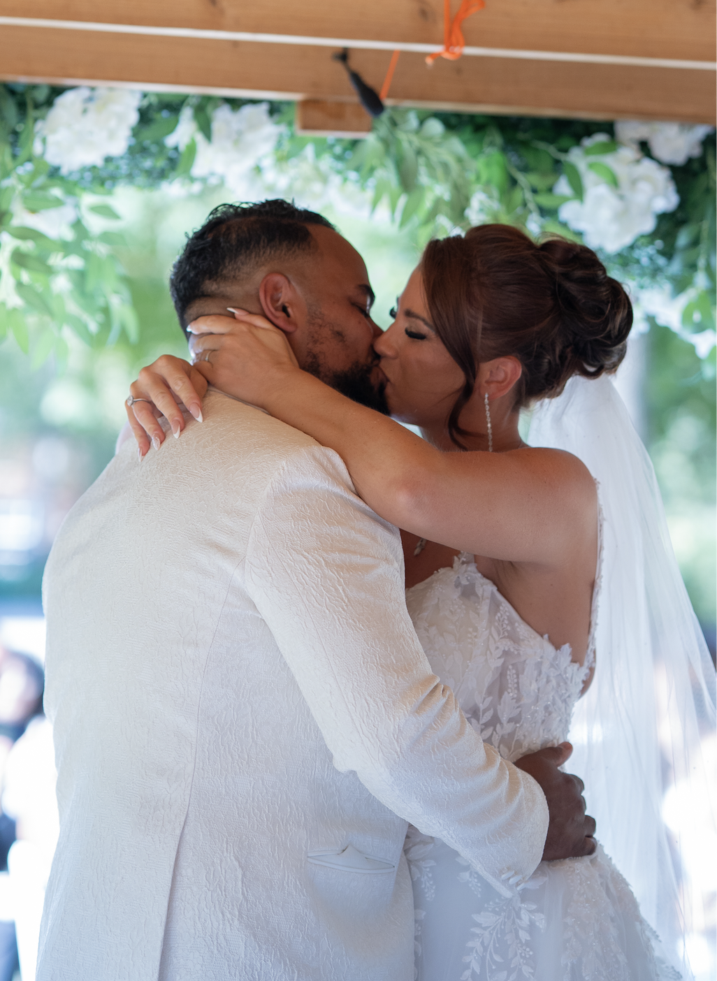 A bride and groom share a kiss at their wedding ceremony, under a floral canopy with greenery and white flowers.