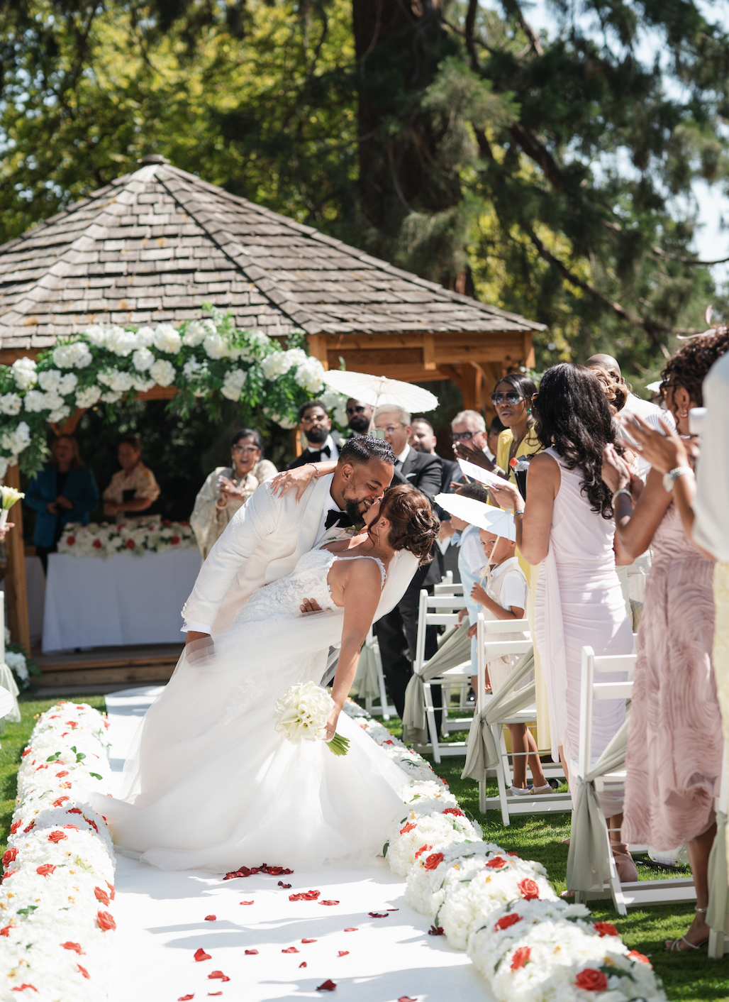 A newlywed couple sharing a kiss at their outdoor wedding ceremony, surrounded by guests clapping and smiling, with floral decorations along the aisle and a wooden gazebo in the background.