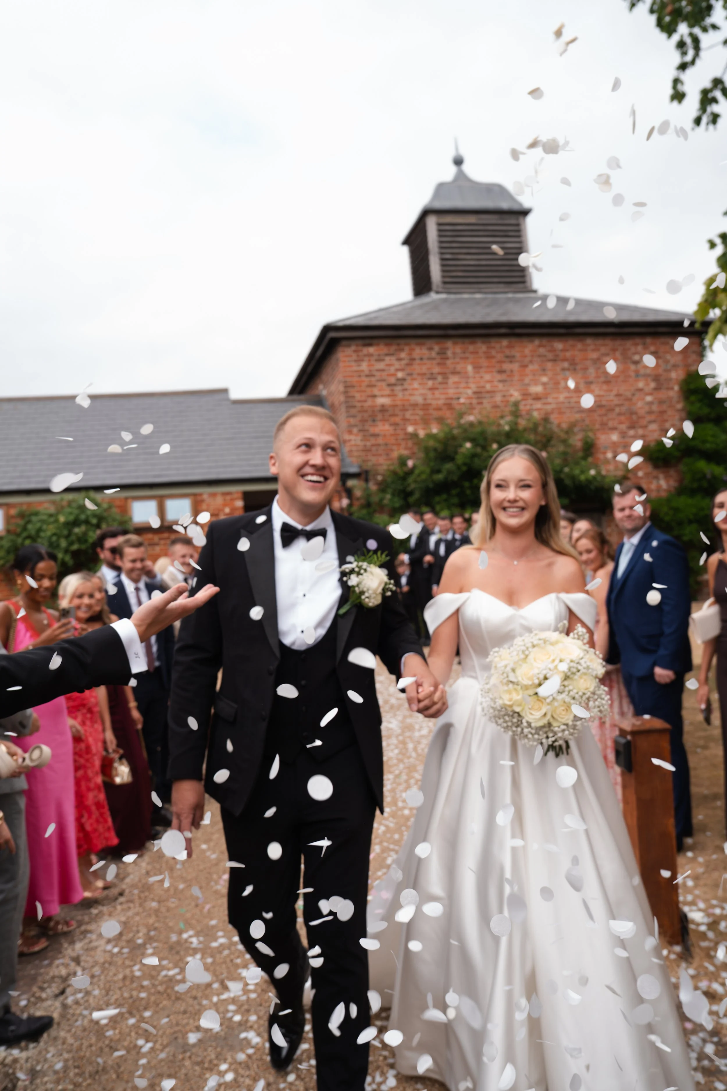 Bride and groom in wedding attire walking hand in hand surrounded by guests throwing confetti outside a brick building with a steeple.