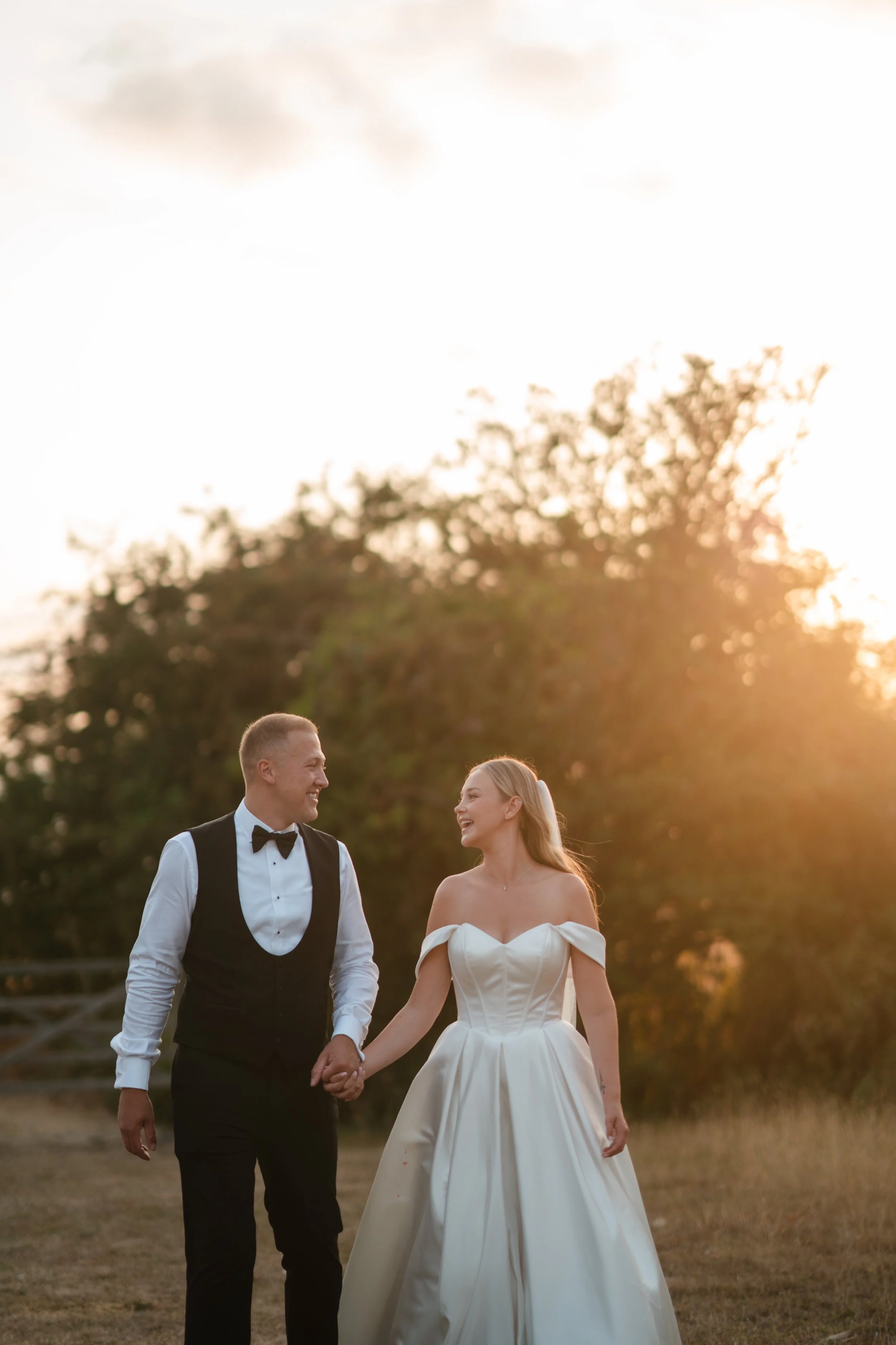 A smiling man and woman holding hands, dressed in wedding attire, walking outdoors during sunset with trees in the background.