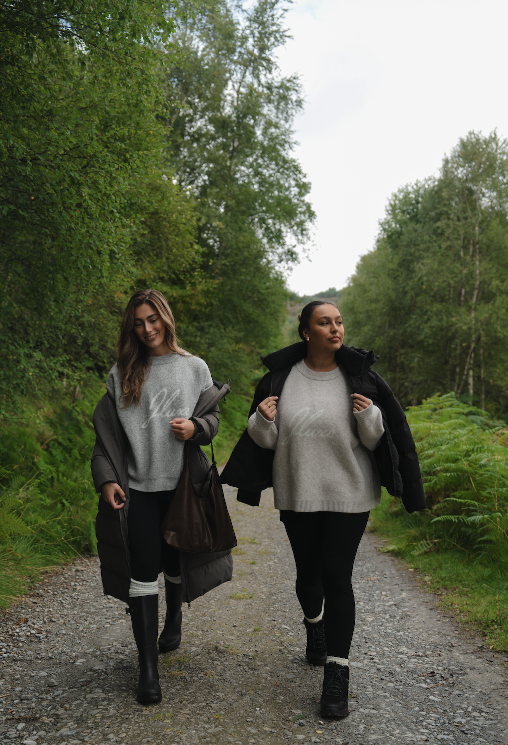 Two women walking along a gravel trail in a lush green forest, both wearing gray sweaters and black jackets, with one smiling and the other looking serious.