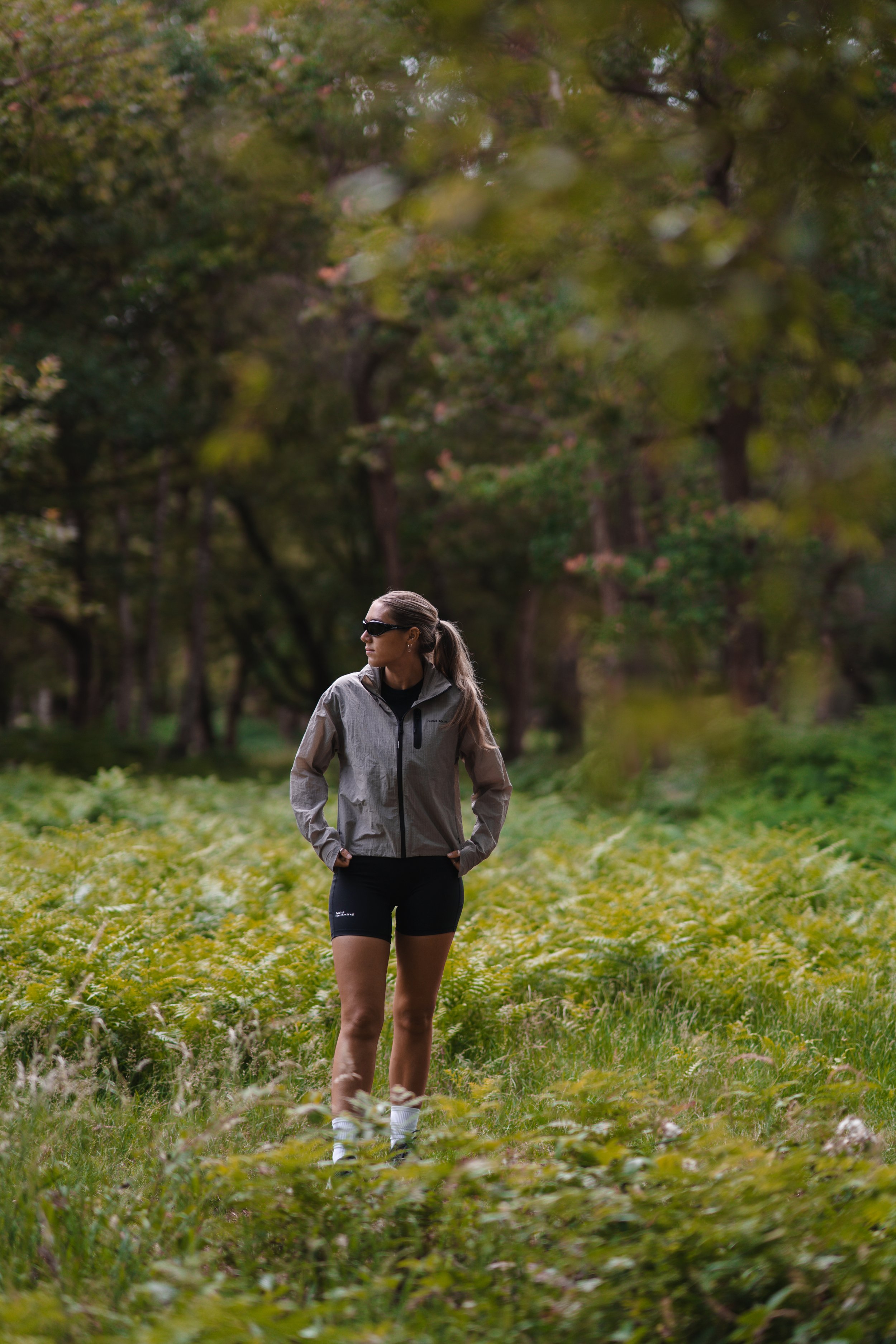 A woman walking in a lush, green park or forest during daytime. She is wearing a light gray jacket, black shorts, white shoes, and sunglasses, with her hair tied back.