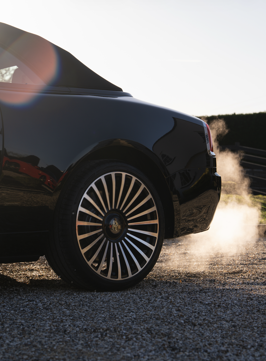 Close-up of a black luxury car with a shiny rim, parked on a gravel surface with steam or smoke coming from the back tire in bright sunlight.