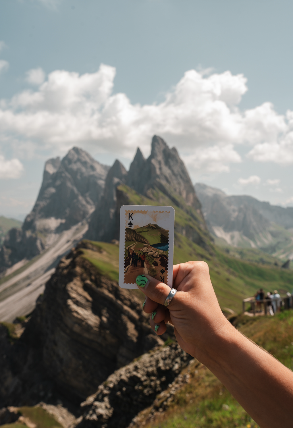 A hand holding a playing card with a scenic illustration of mountains, a lake, and hikers, against a background of towering mountain peaks and a cloudy sky.