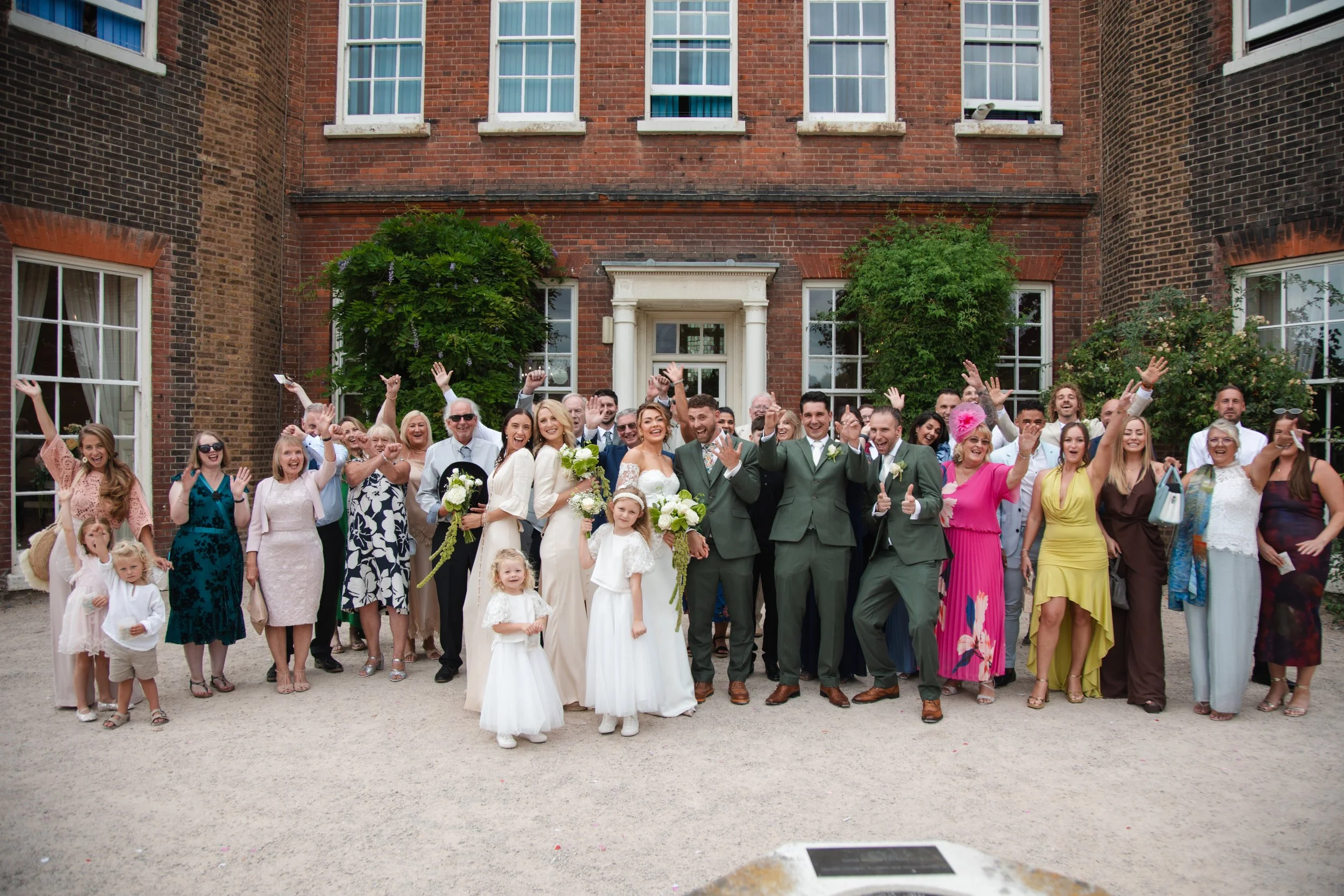 Group of wedding guests and couple standing in front of a brick building, celebrating and smiling, with some raising their hands and others holding bouquets.