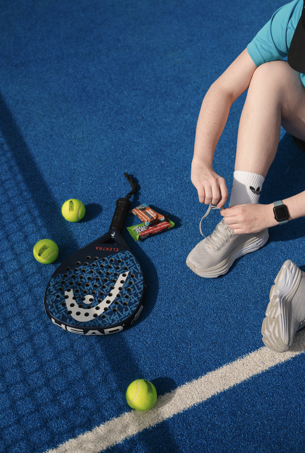 Person tying shoelaces on white athletic shoes on a blue court, with tennis balls and a paddle nearby.