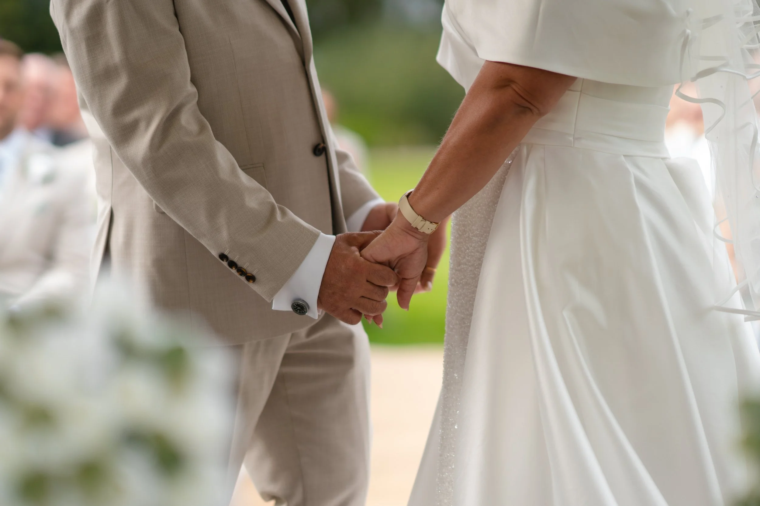 Close-up of a bride and groom holding hands during a wedding ceremony, with focus on their clasped hands, wedding attire, and a blurred outdoor setting in the background.