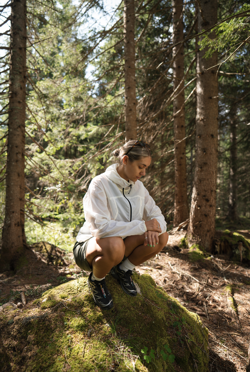 A woman crouching on a moss-covered rock in a dense forest with tall trees and sunlight filtering through the branches.