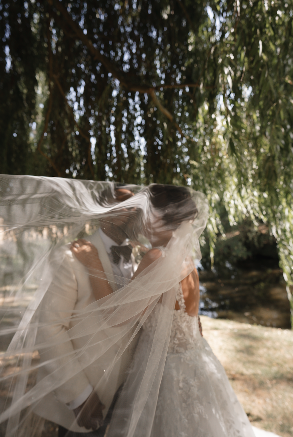 A bride and groom standing close together outdoors, with the groom gently covering the bride's face with a sheer veil. The bride is wearing a wedding dress with lace details, and the groom is in a light-colored suit with a bow tie. The background features trees and sunlight.
