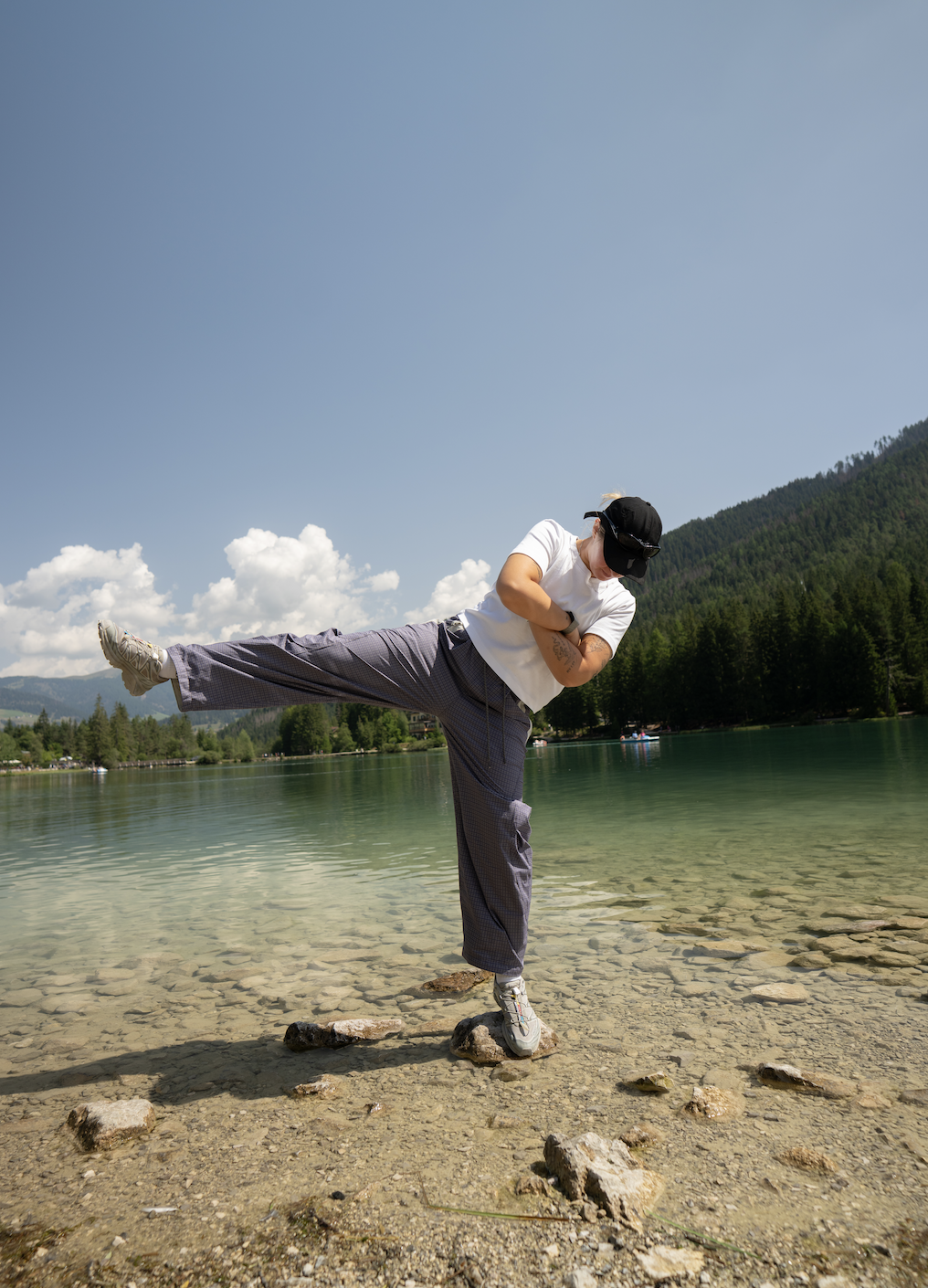 Person performing a martial arts move or balancing pose barefoot on rocks at a lakeshore with mountains and trees in the background on a sunny day.