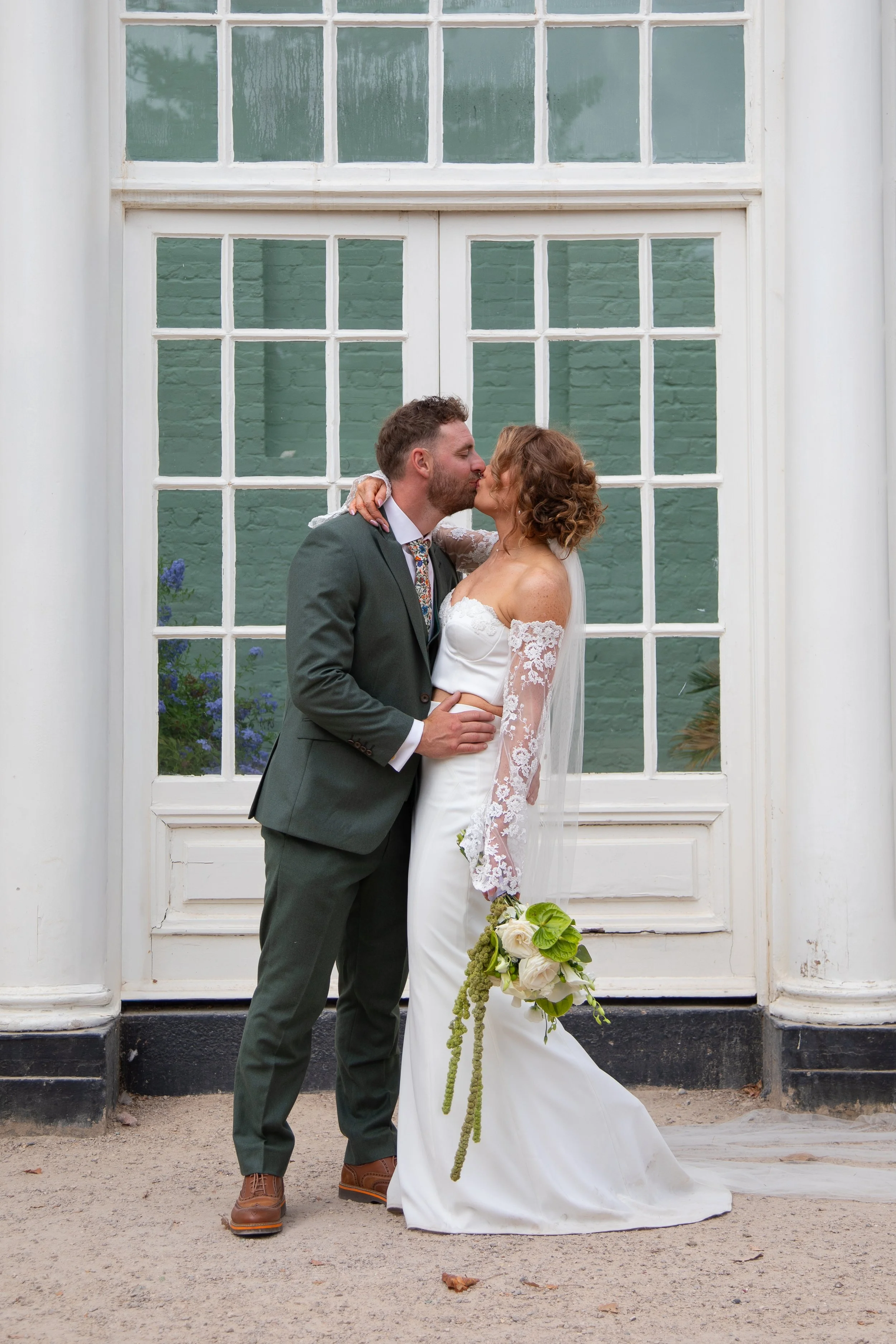A bride and groom share a kiss outside a large window with white framing. The bride wears a white lace wedding dress and holds a bouquet of white and green flowers. The groom wears a gray suit with a colorful tie and brown shoes.