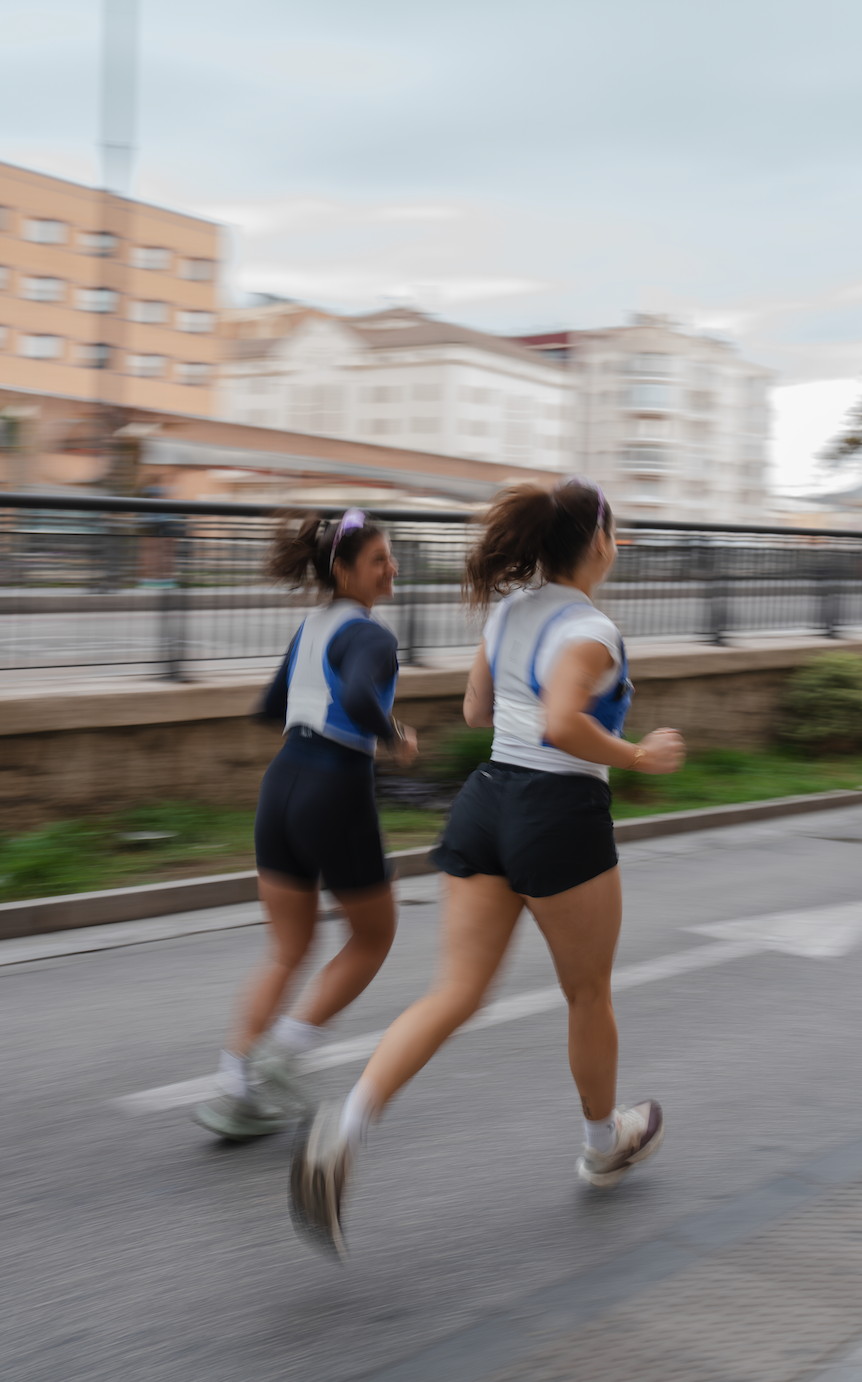 Two women running on the street, blurred motion indicating speed, background of residential buildings and a cloudy sky.