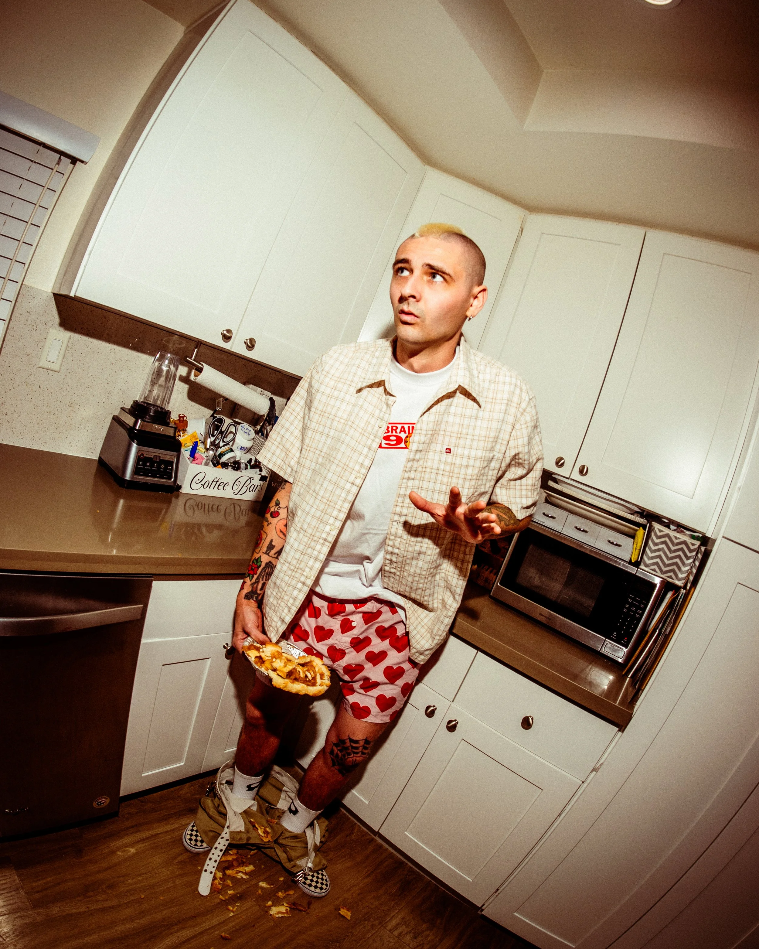 A man with tattoos wearing heart-patterned boxer shorts and checkered shoes standing in a kitchen with pantry over, holding a pie, with spilled pie and crumbs on the floor.