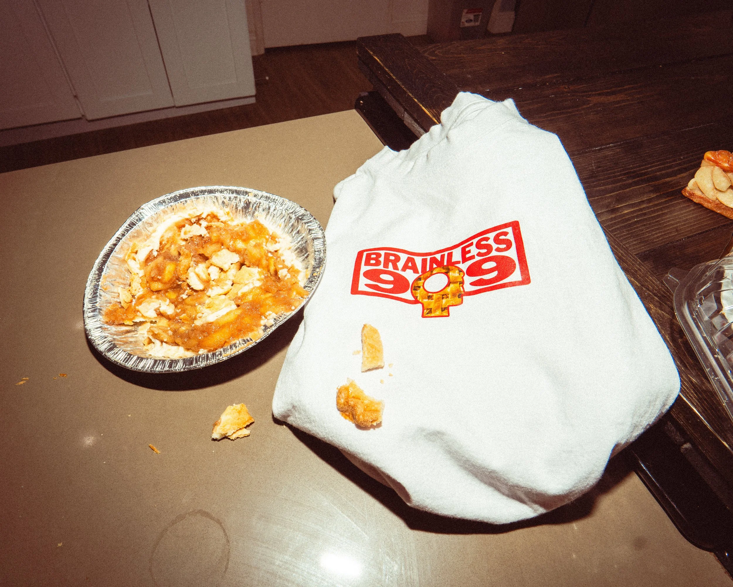 A partially eaten apple pie in a foil container next to a white cloth bag with red text and graphics that read 'BRAINLESS 99', on a brown table.