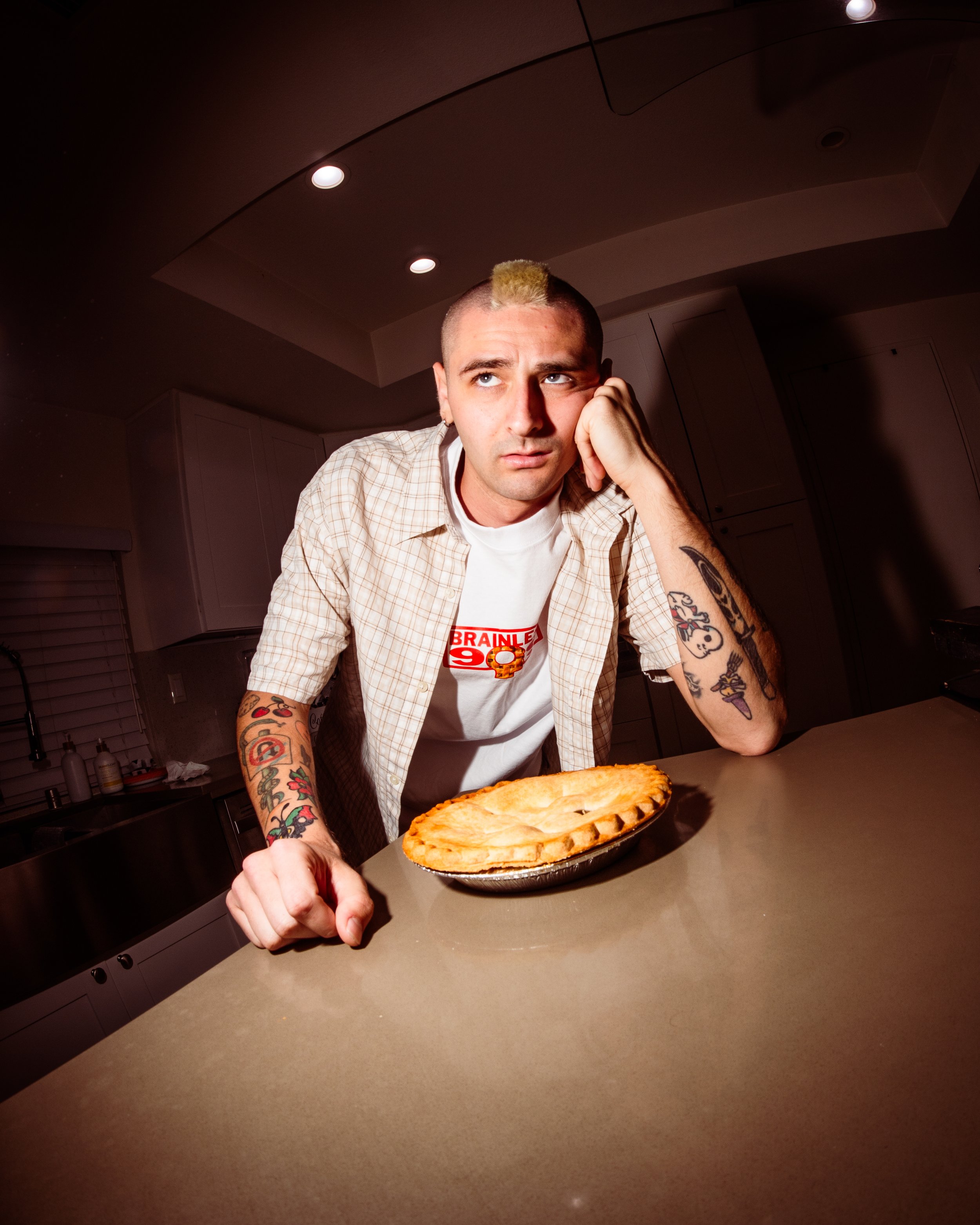 A man with tattoos, wearing a plaid shirt and a white t-shirt with red and yellow text, sitting at a kitchen counter with a pie in front of him, looking bored or annoyed.