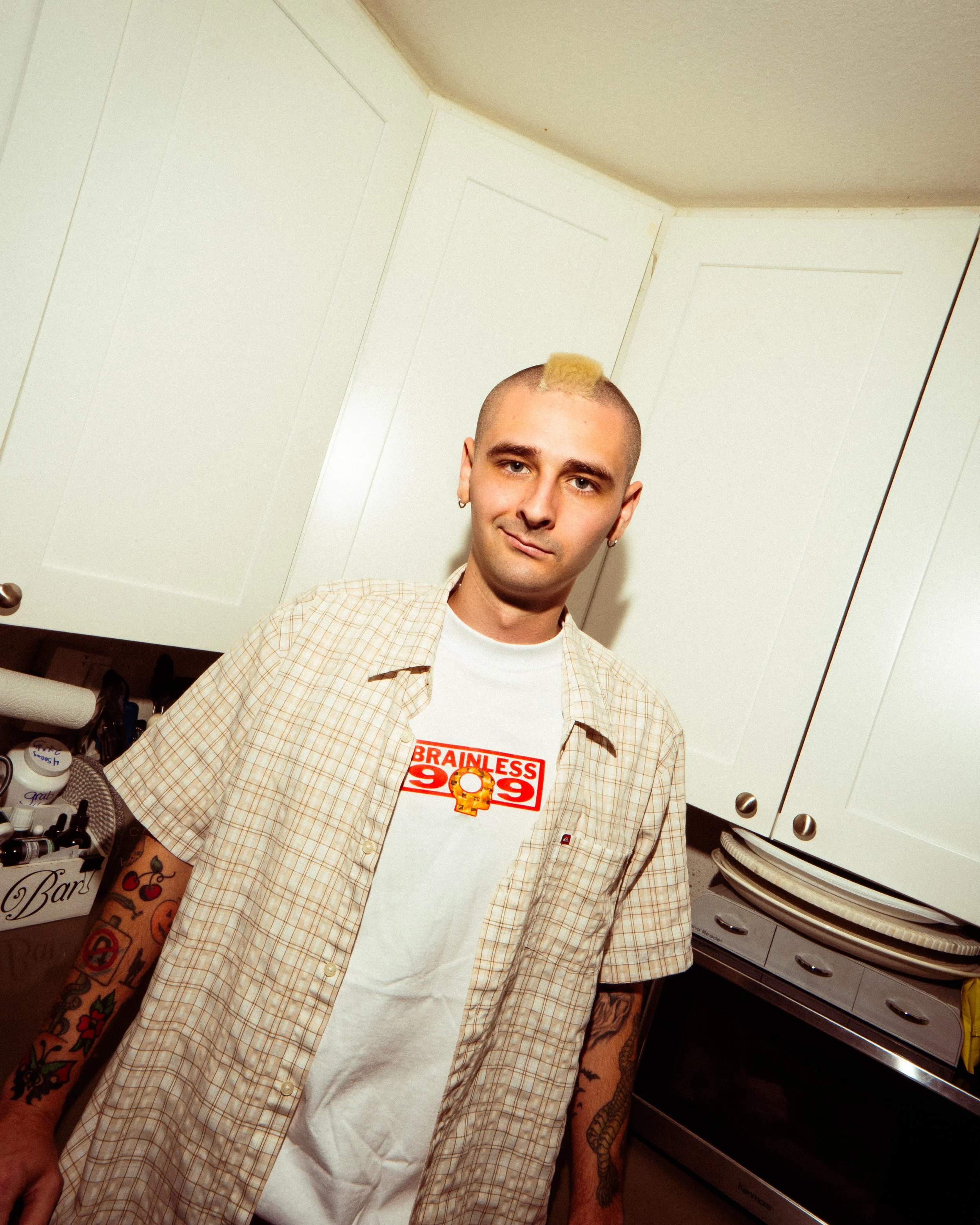 A young man with a shaved head and a yellow mohawk hairstyle, wearing a beige checkered shirt over a white T-shirt with red text and an illustration, standing in a kitchen with white cabinets and a black stove.