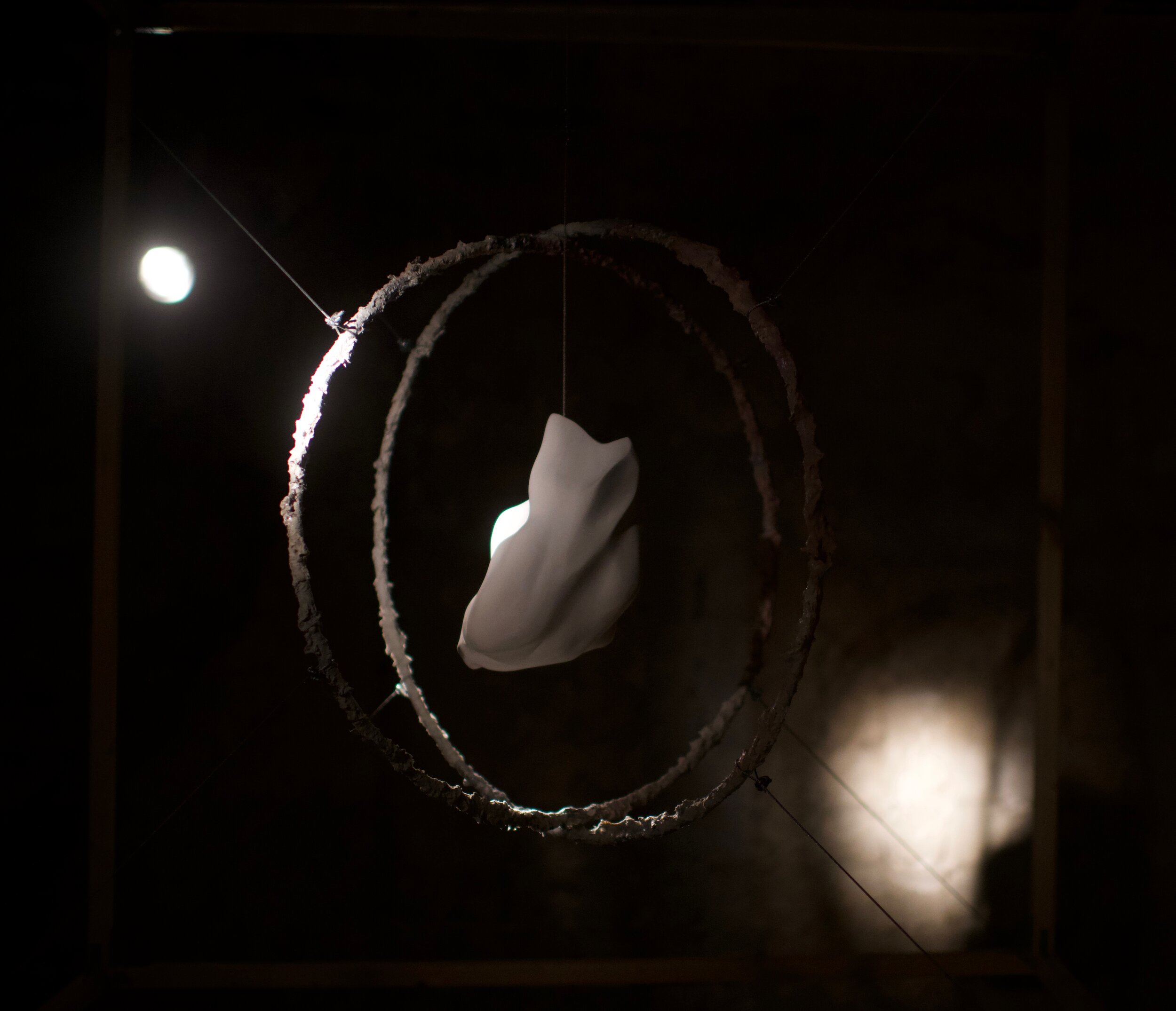 Abstract art installation with a white sculptural form suspended within two rustic metal hoops, illuminated in a dark setting.