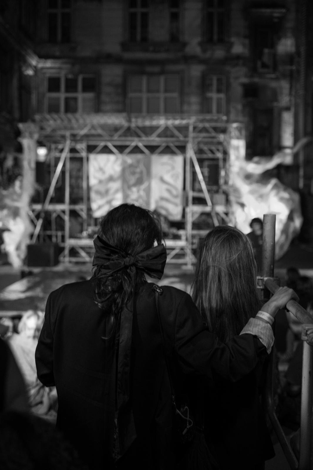 Two people in formal attire, one with a large bow headband, standing in front of a stage with a backdrop, in an urban outdoor setting at night.