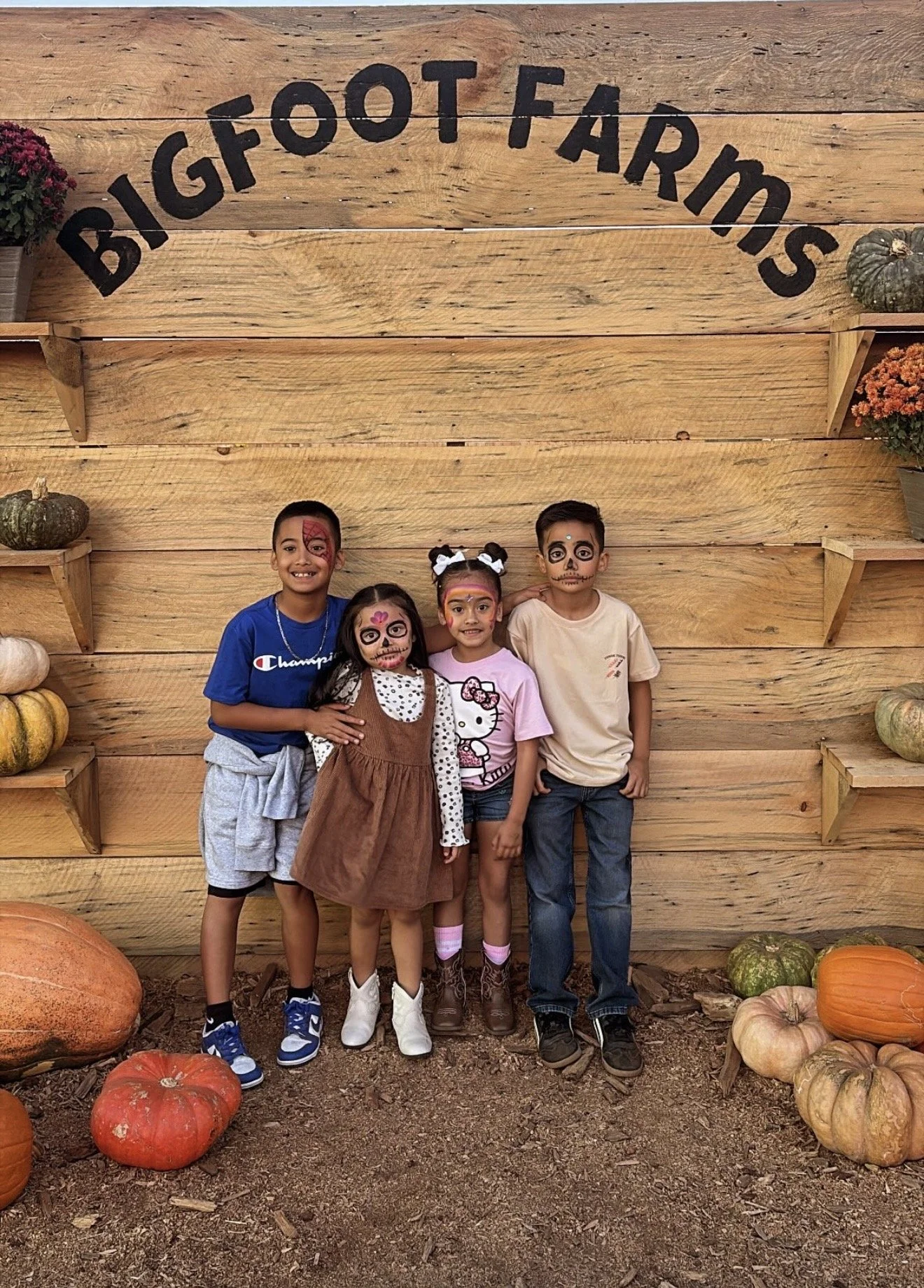 Four children with face paint in front of a wooden wall with pumpkins and flowers, during fall.