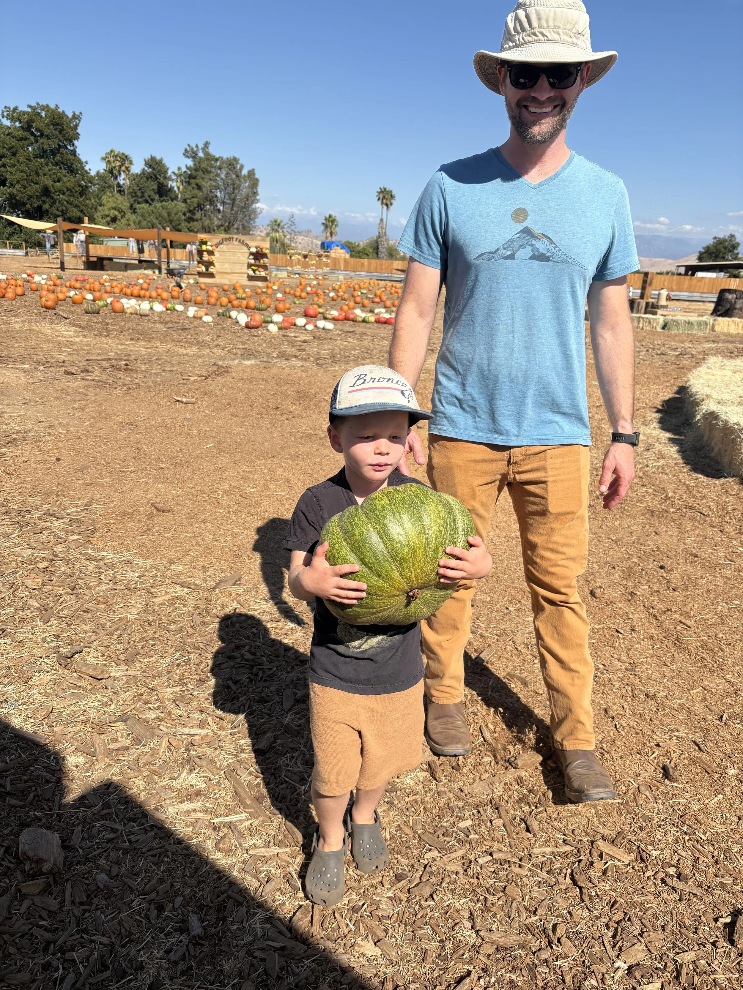 A man and a boy at a pumpkin patch, with the boy holding a large green pumpkin. The man wears a hat, sunglasses, a blue t-shirt, and brown pants, while the boy wears a cap, black shirt, beige shorts, and Crocs shoes.