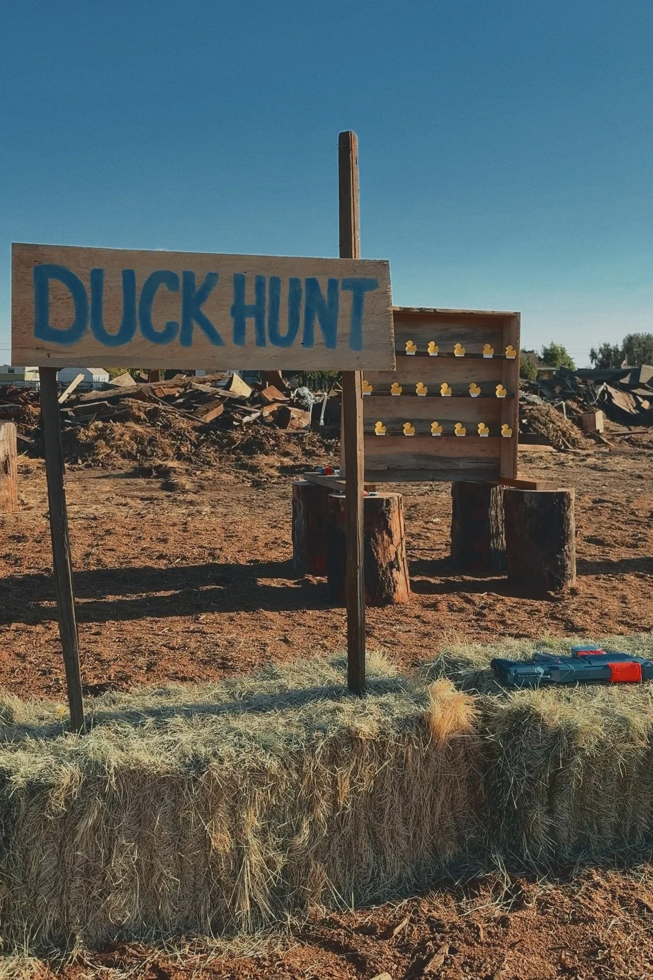 A sign that says 'Duck Hunt' and a vertical wooden rack with duck-shaped targets, set up outside on a dirt field with clear sky.