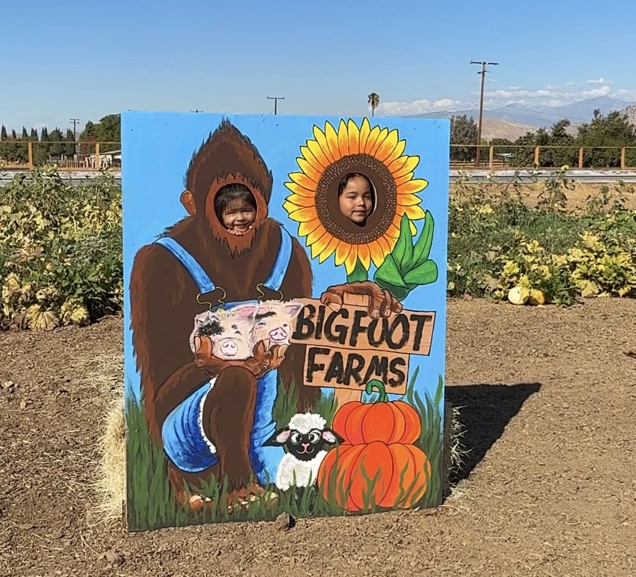 Children peeking through holes in a painted Bigfoot Farms display, featuring a Bigfoot figure holding piglets, a sunflower, pumpkins, and a sheep, with a farm background and clear skies.
