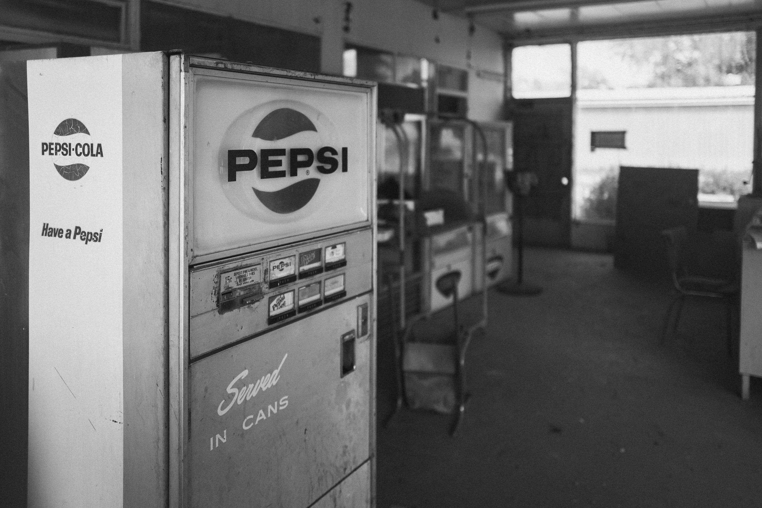 A vintage Pepsi-Cola vending machine inside an abandoned store, with sunlight streaming through the doorway.