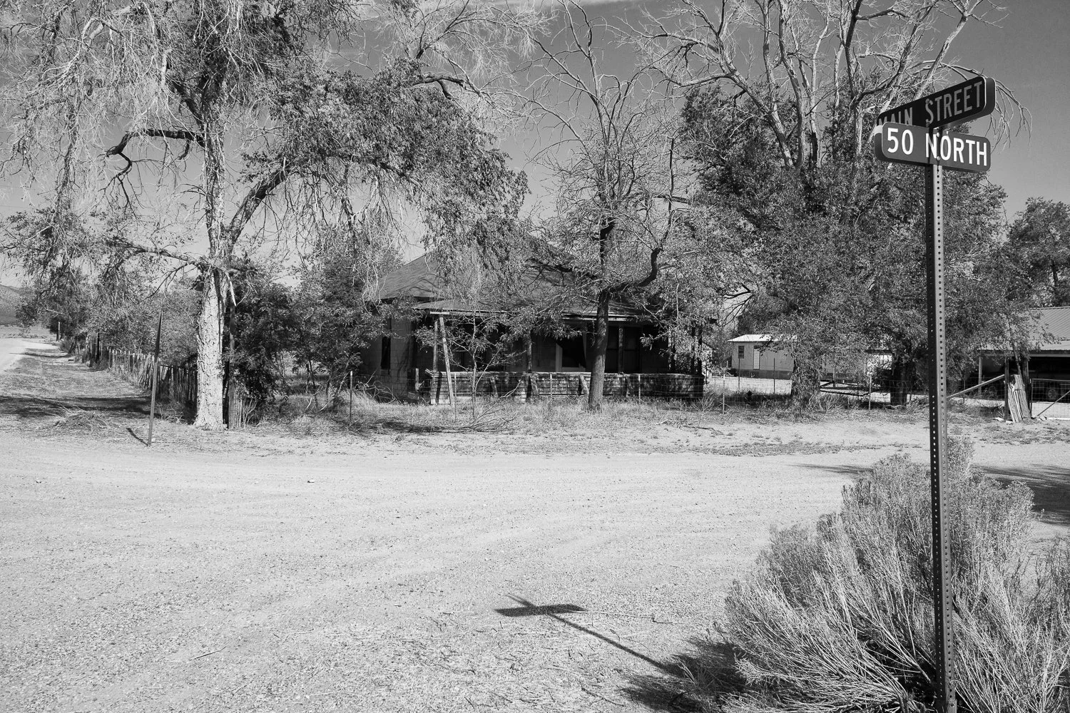 A leaning street sign for 50 North and Main Street beside a dirt road and an abandoned house.