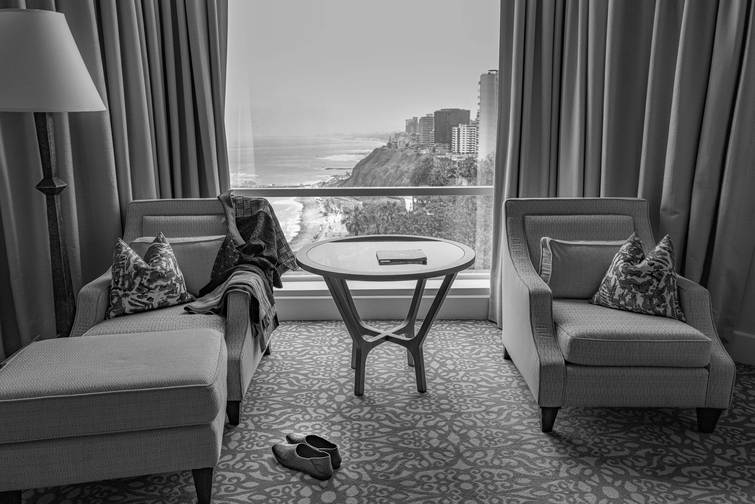 A chaise and armchair by a window overlooking a coastal city skyline and beach.