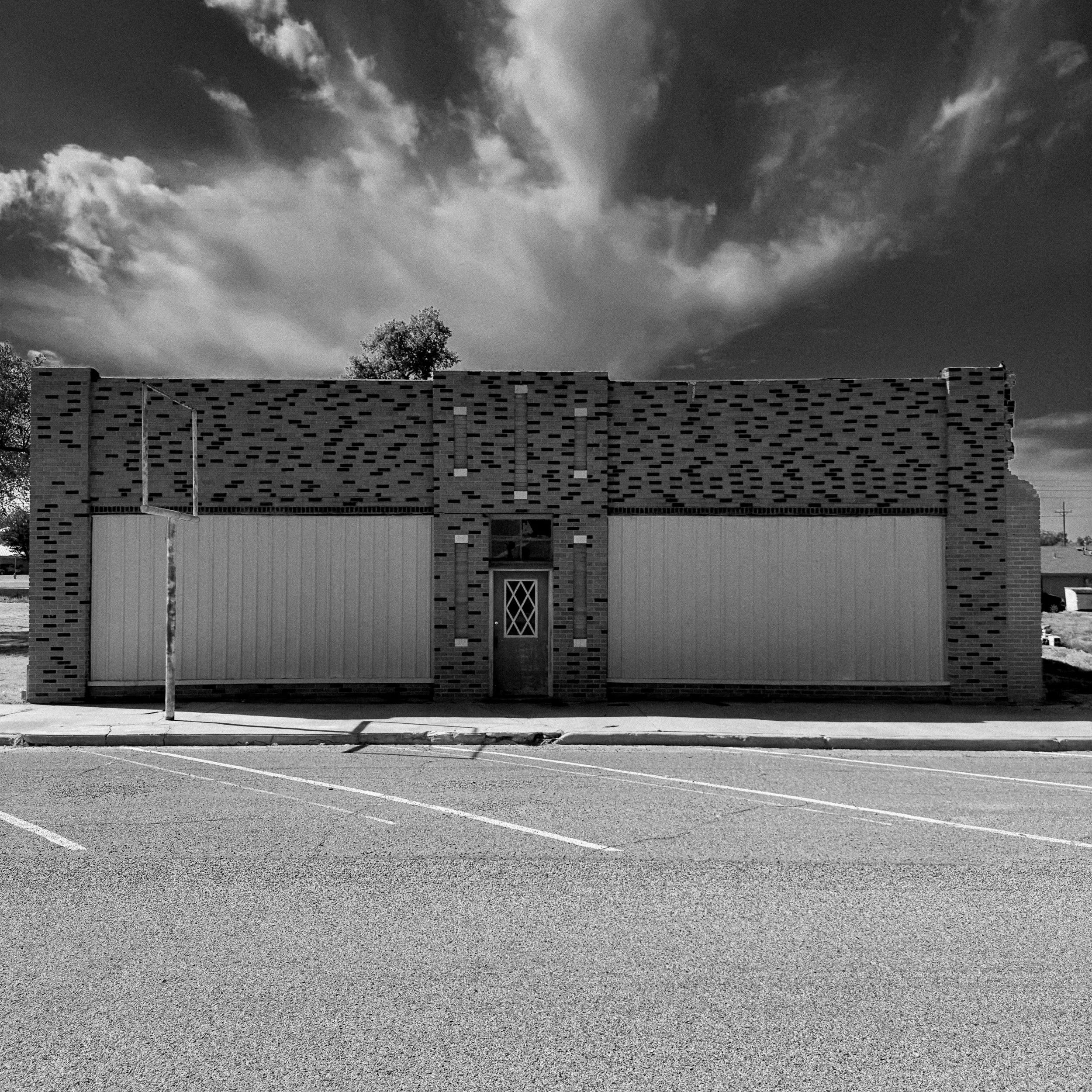 A patterned brick storefront with boarded panels and a diamond-paned door beneath dramatic clouds.