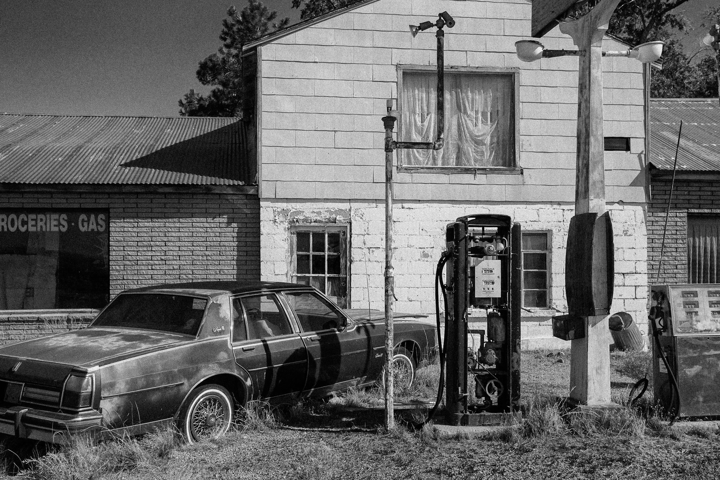 A rusted vintage sedan parked beside two old-fashioned gas pumps in front of a boarded general store.